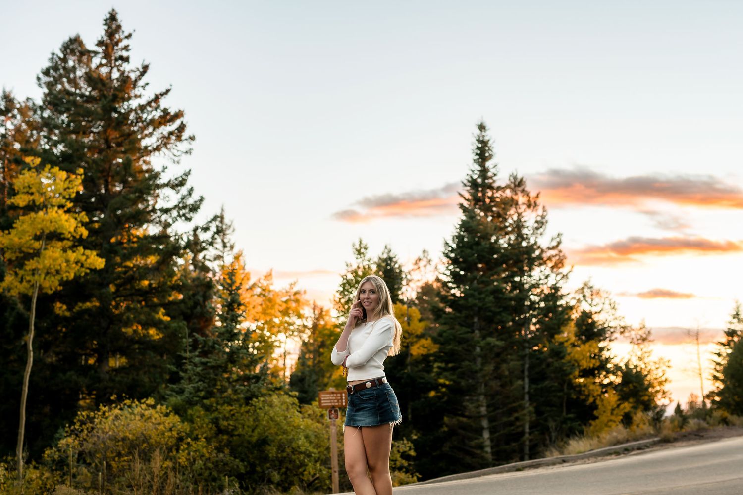 Young blonde woman in white top and denim shorts poses on a road surrounded by autumn trees at sunset.