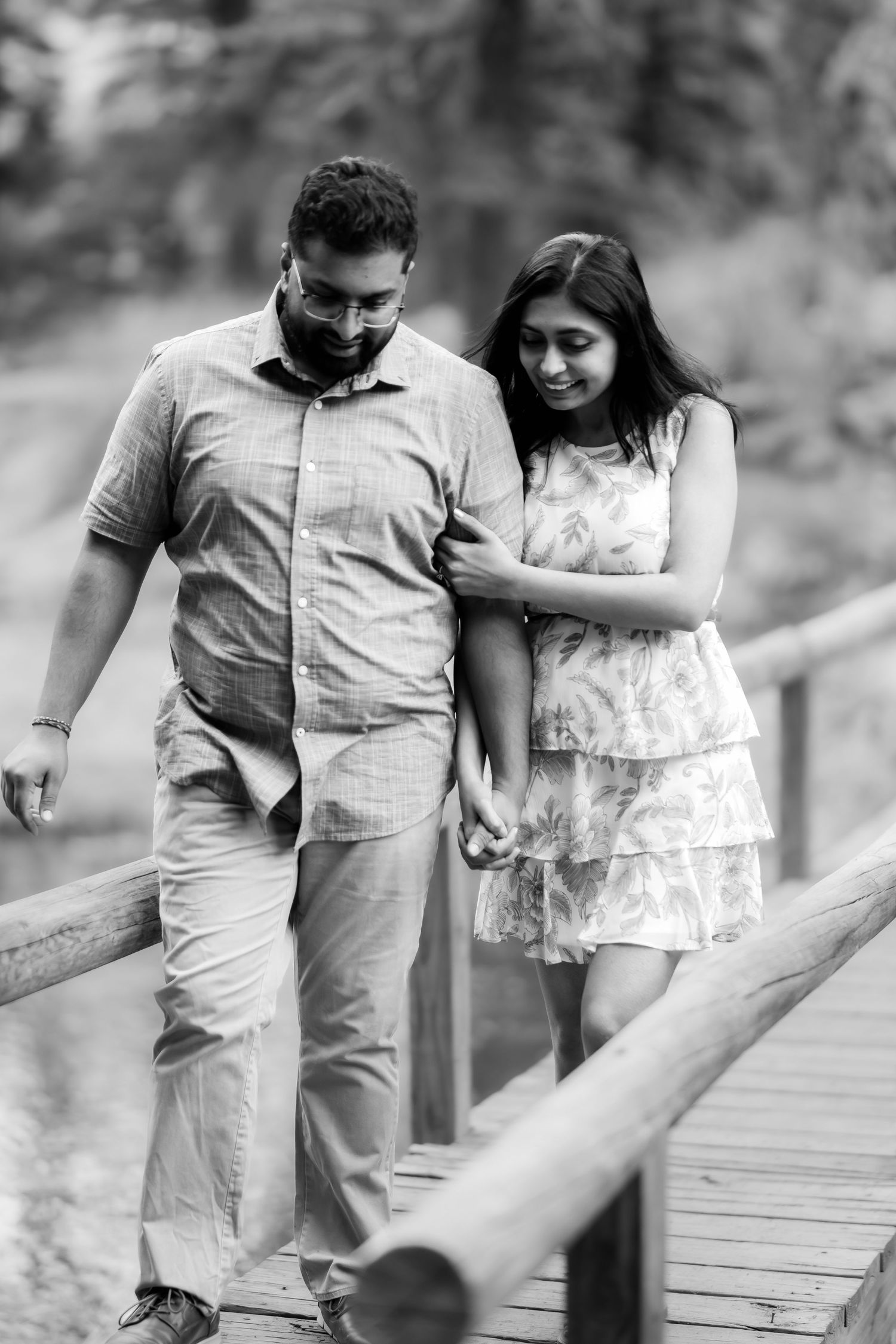 A black and white photo of a couple walking hand in hand along a wooden bridge in nature.