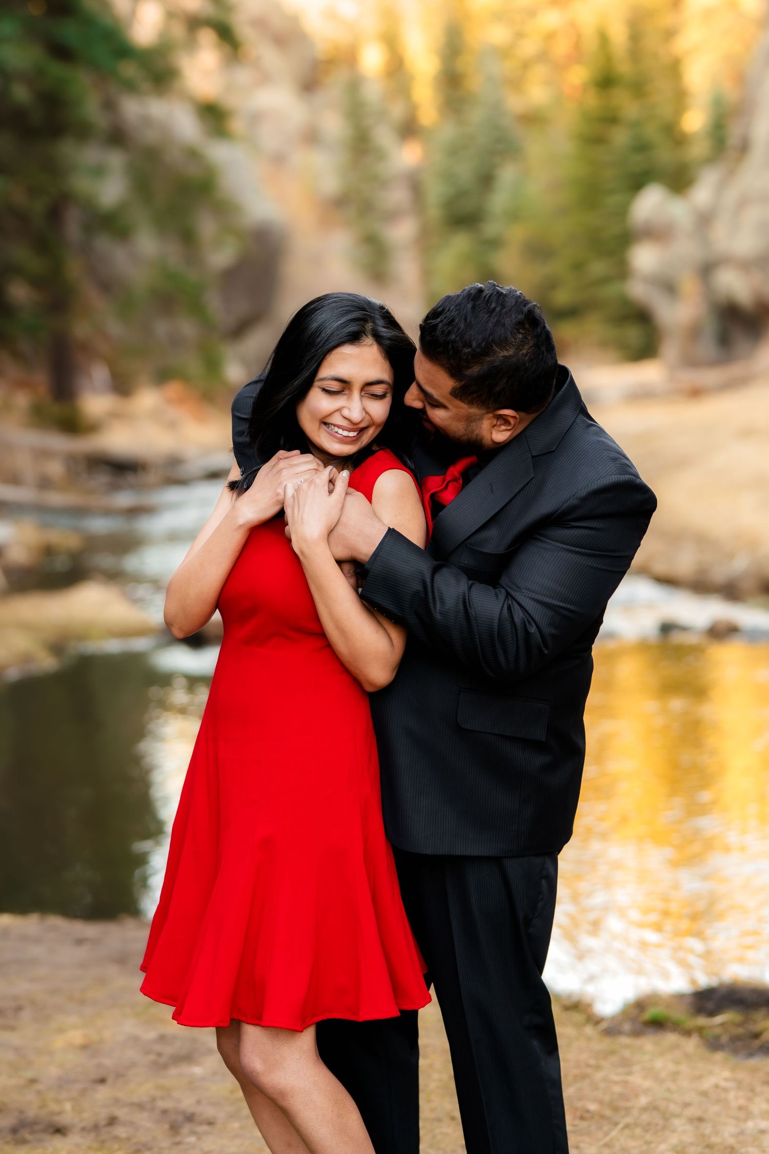 A romantic couple embraces by a scenic lake at sunset, with her wearing a red dress and him in a black suit.