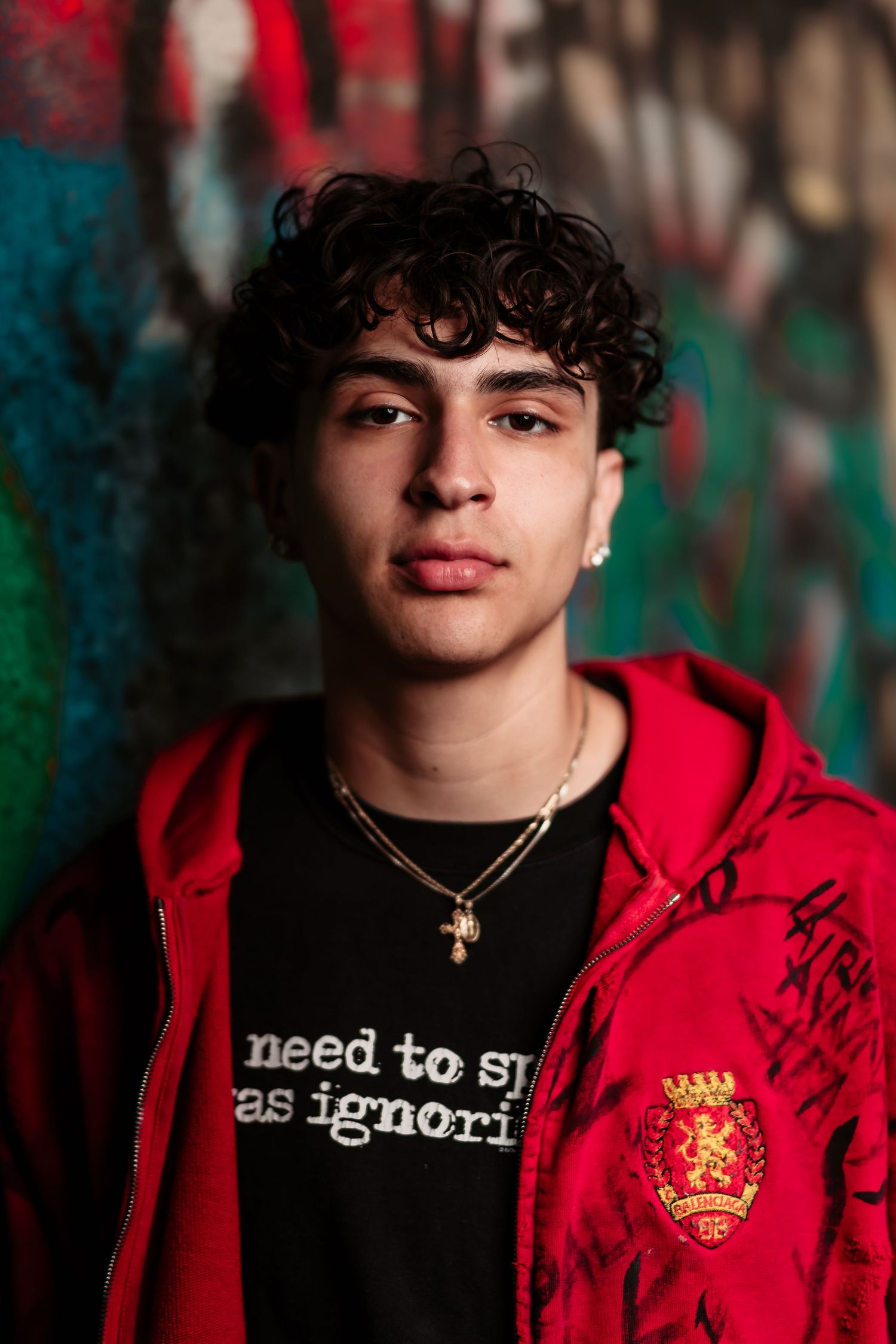 Portrait of someone wearing a red hoodie and cross necklace against colorful graffiti background.