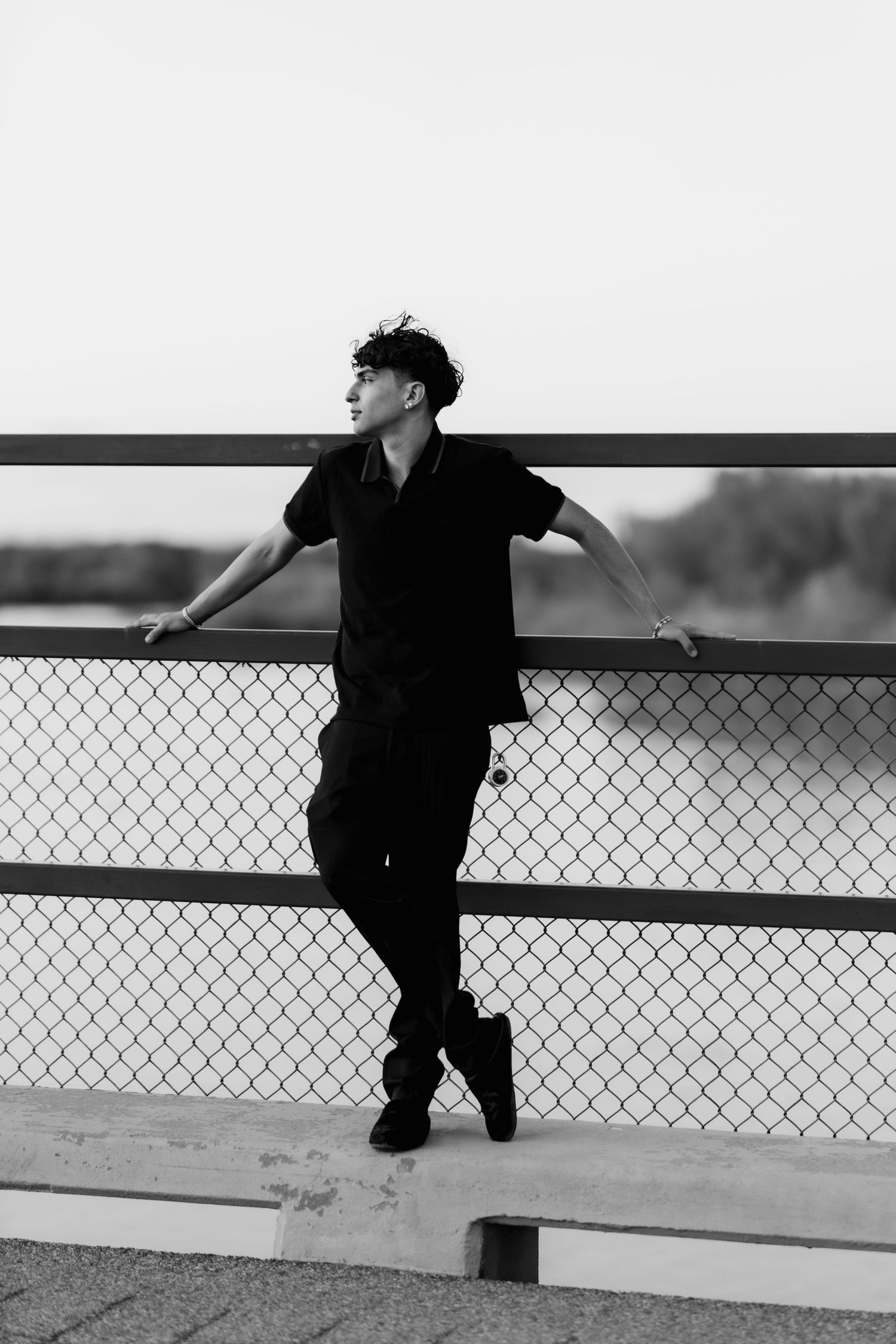 A silhouette of a person in black attire leans against a chain link fence in an artistic black and white composition.