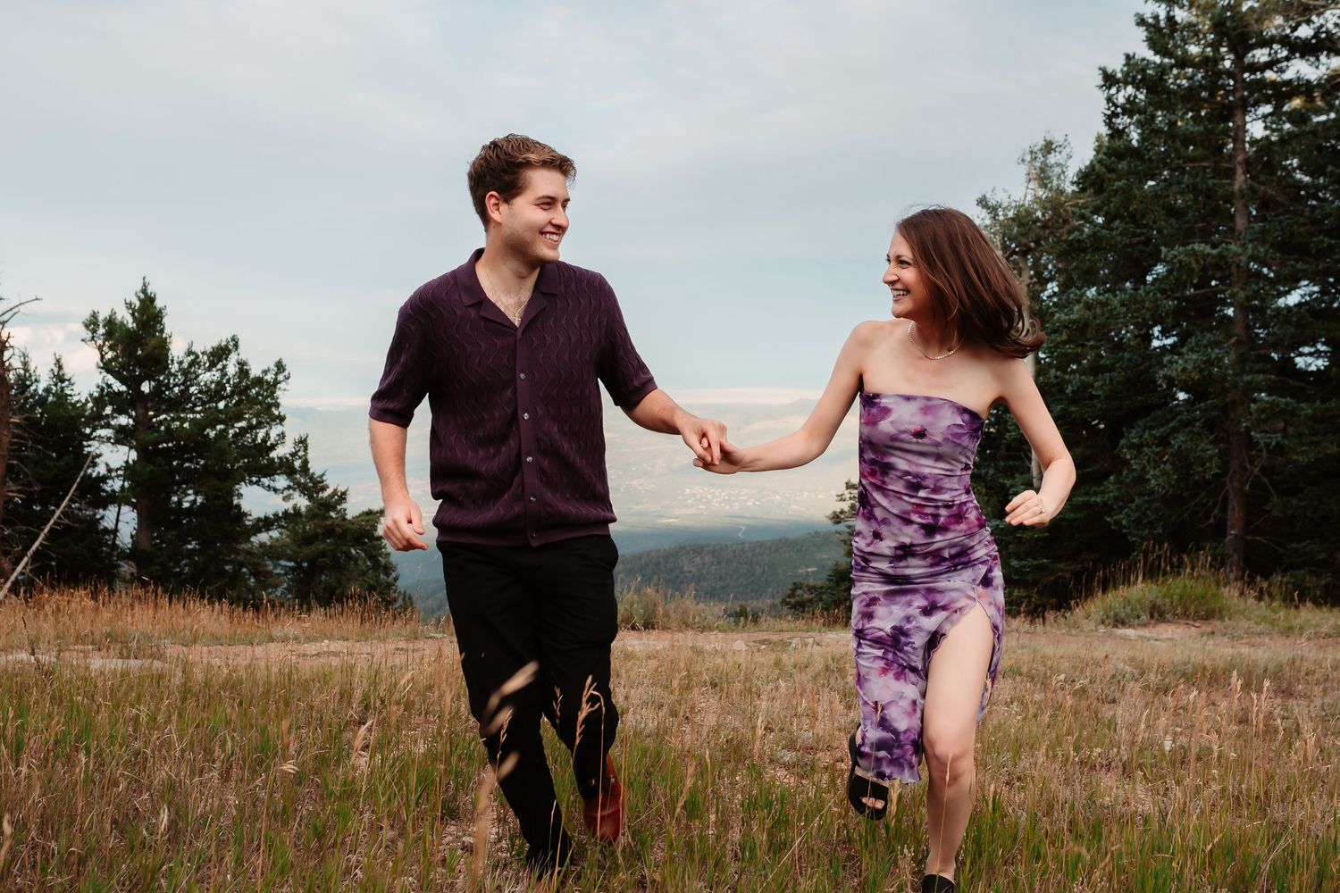 Dancing couple enjoys playful moment in mountain meadow with evergreen trees in background.