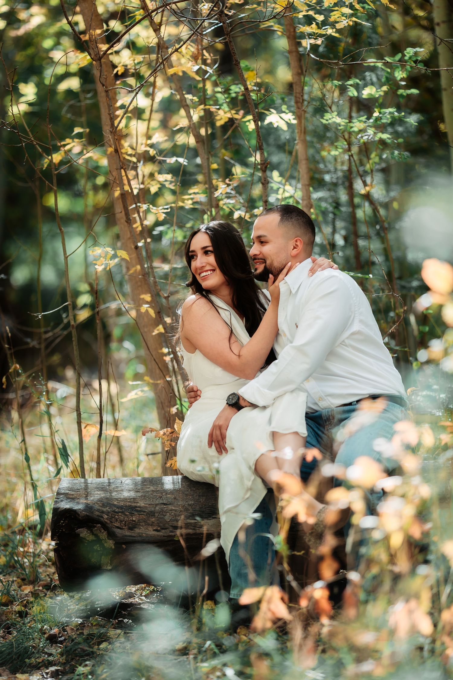 A couple in white outfits share intimate moments while sitting together among leafy trees and dappled sunlight.