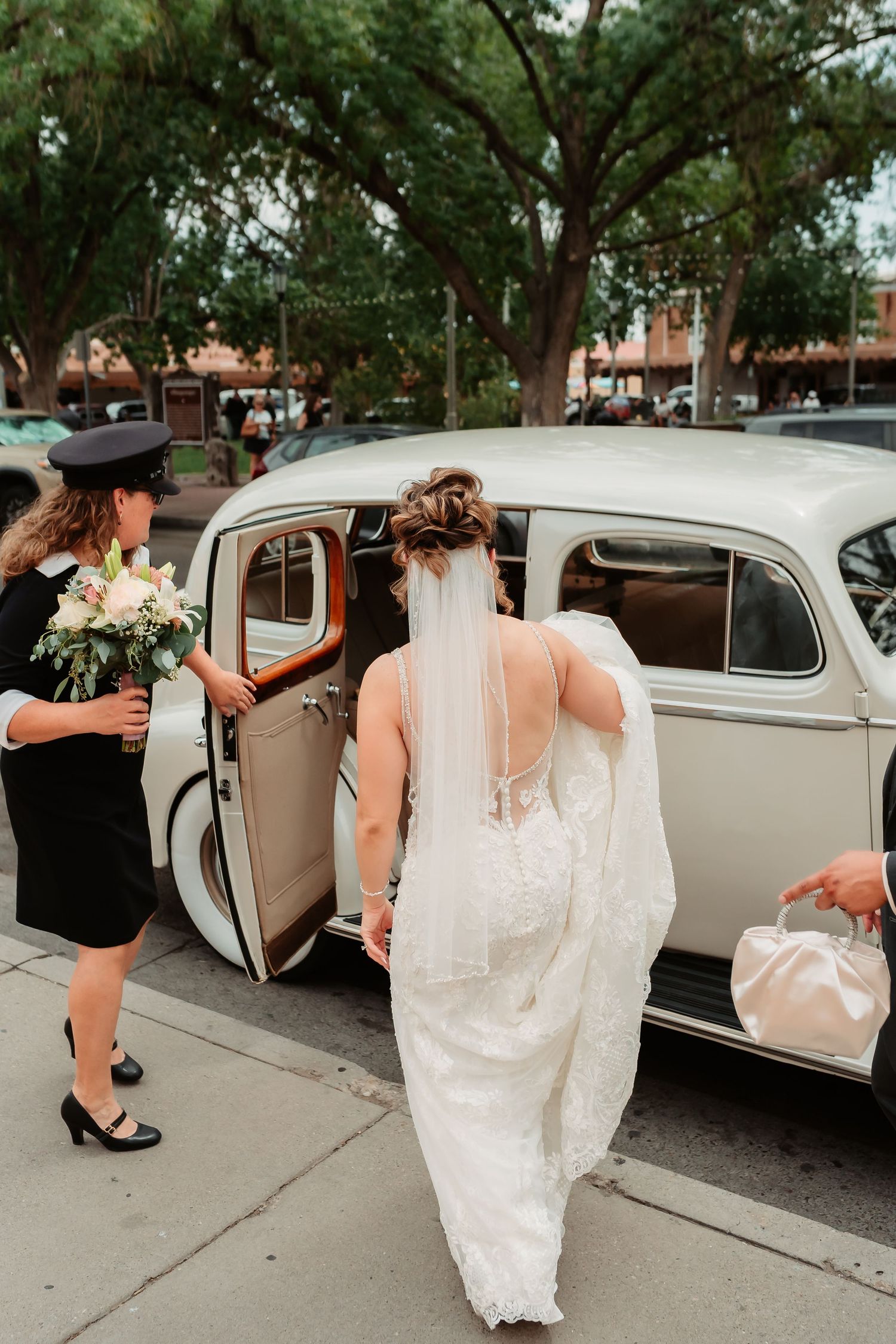Wedding party helps bride into vintage cream-colored classic car on tree-lined street.