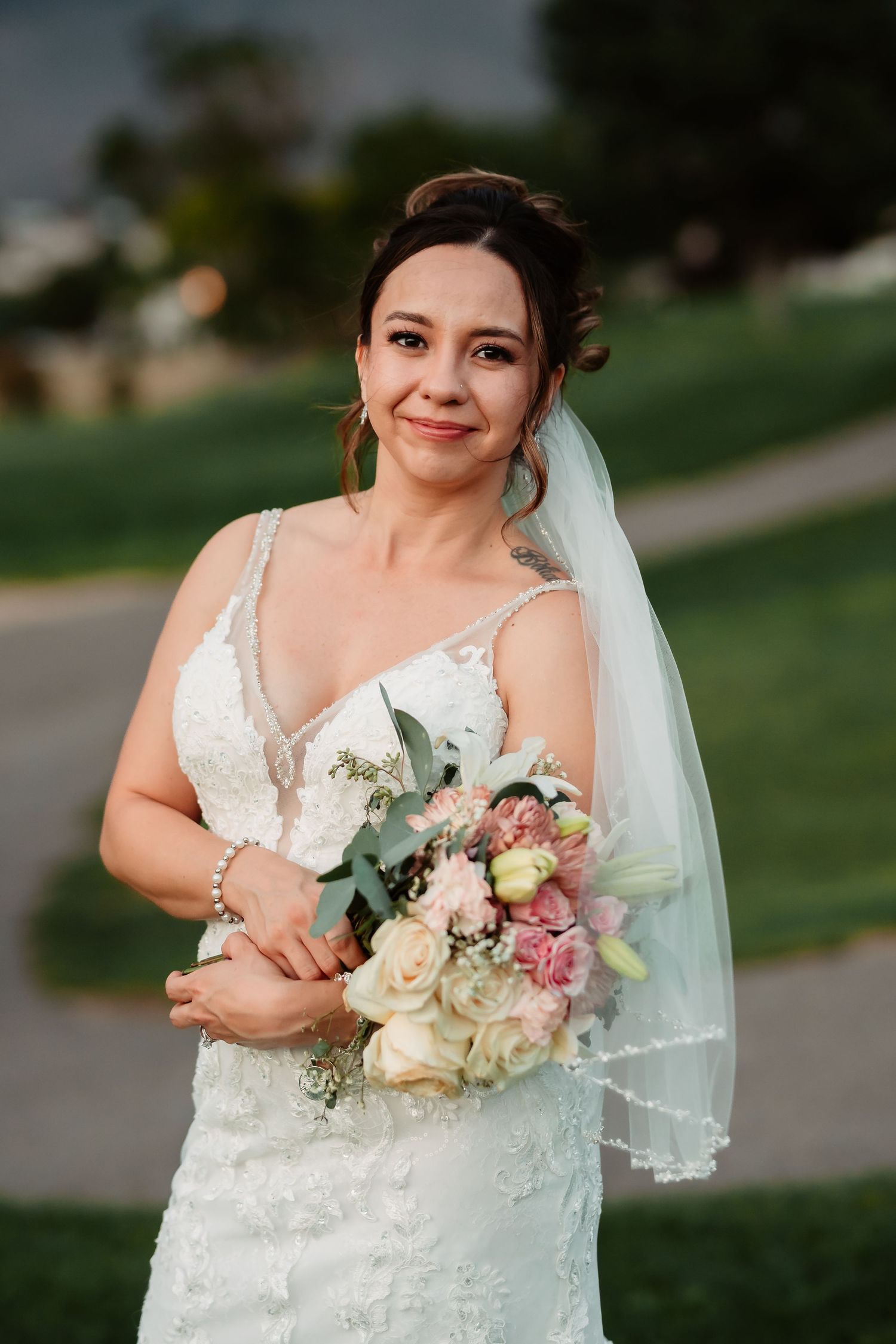 A bride wearing a sleeveless lace gown and veil smiles while holding a bouquet of pink and white flowers.