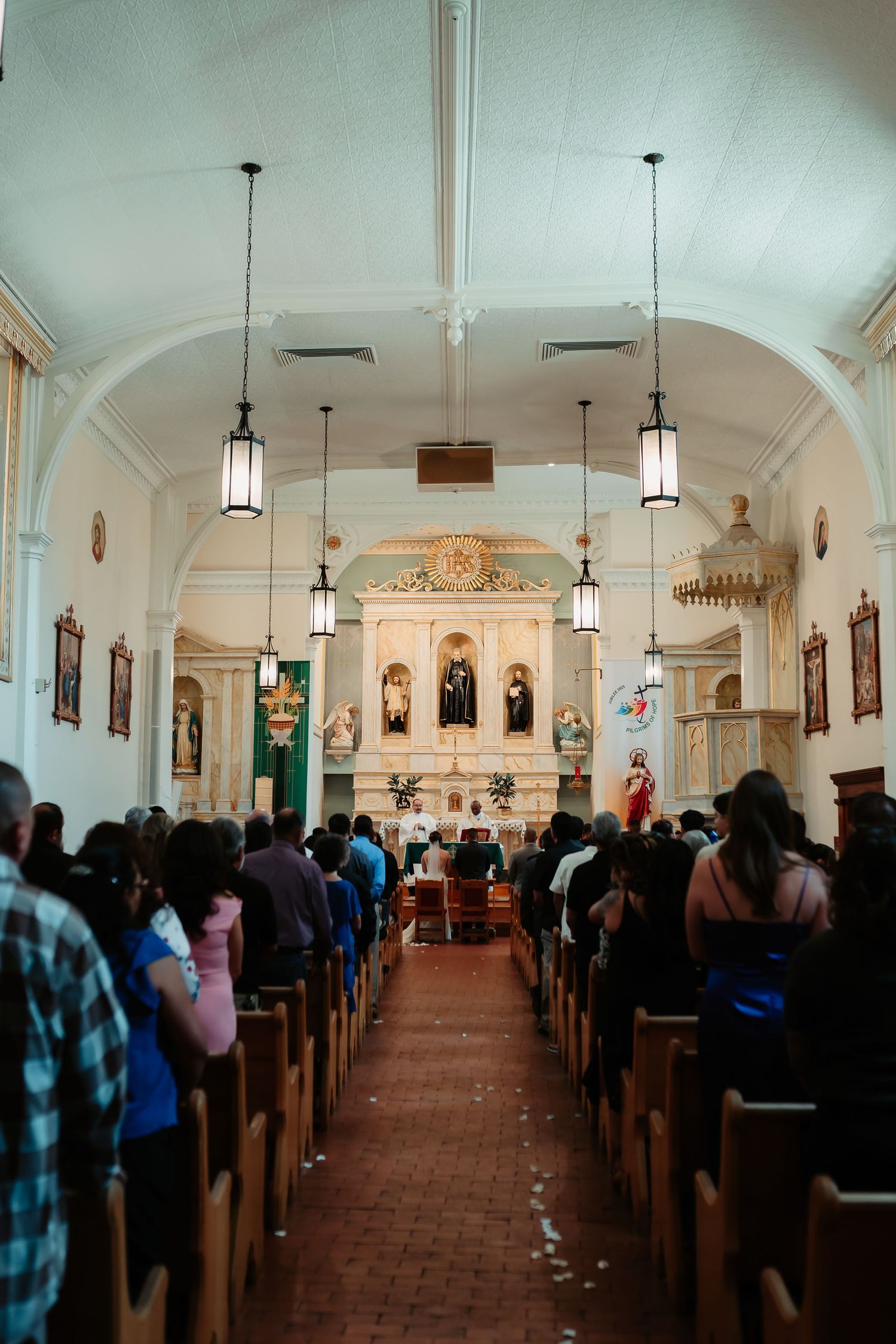 Interior of a traditional Catholic church with arched ceilings and pendant lights during a wedding ceremony.