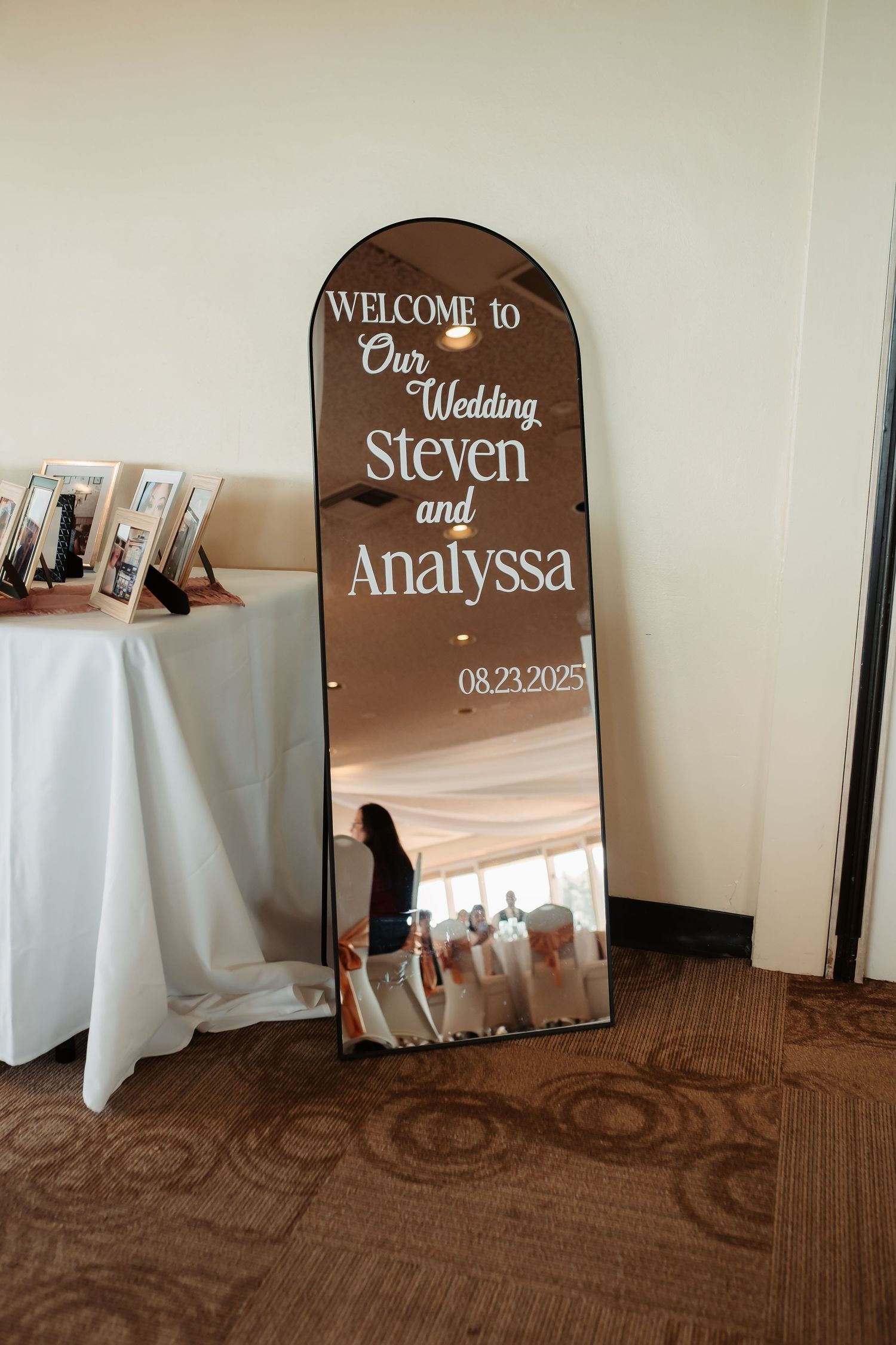 Wedding welcome sign displays mirror with elegant brown wooden frame and text placed on white linen covered tables.