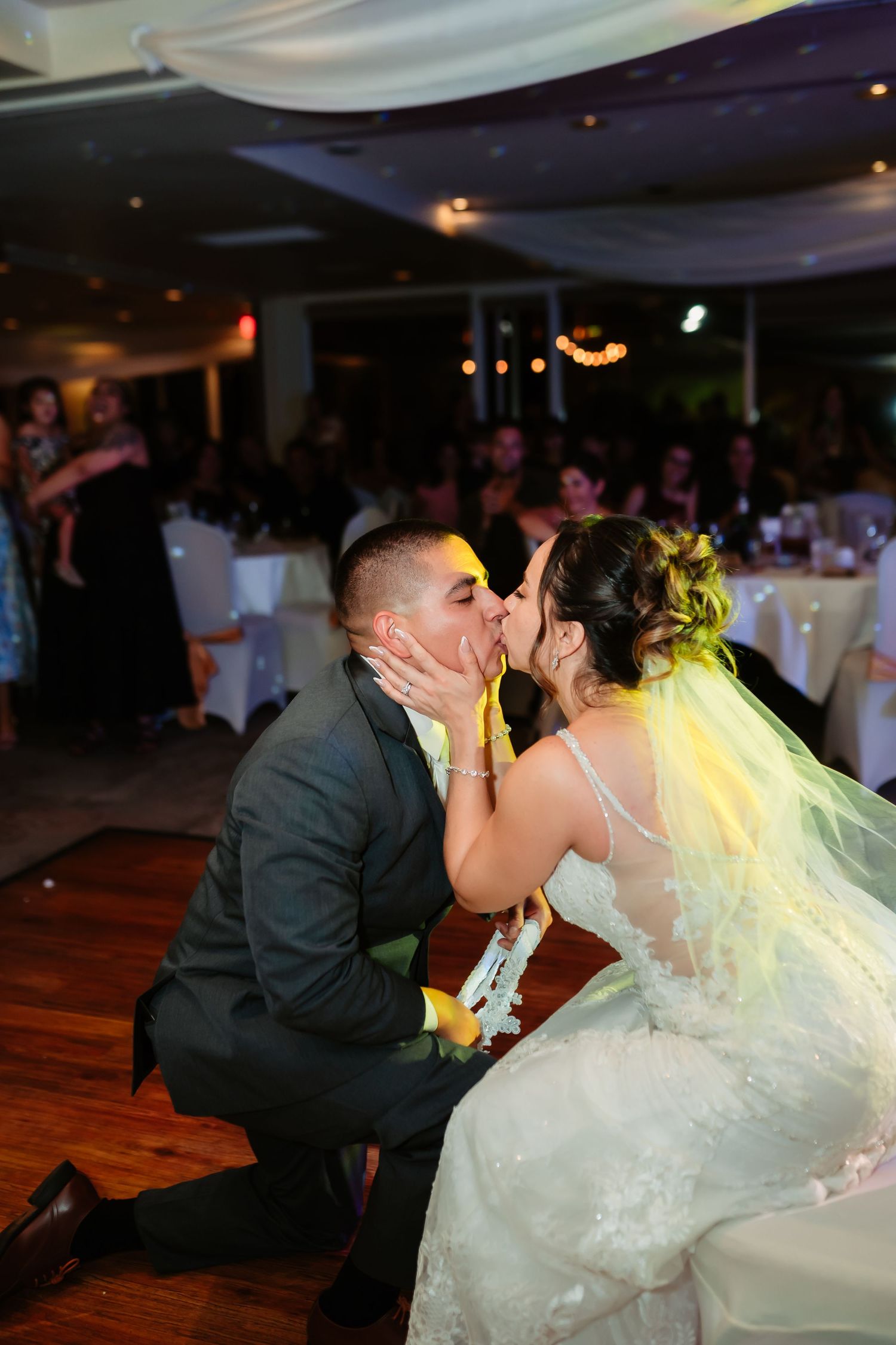 Newlyweds share a romantic kiss during their wedding reception on a wooden dance floor with decorative lighting.
