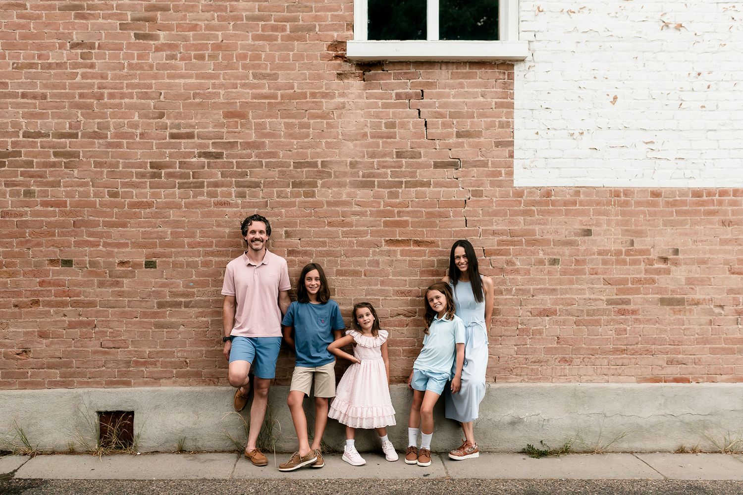 Family of five smiling against a brick wall outdoors, wearing casual pastel outfits on a sunny day.