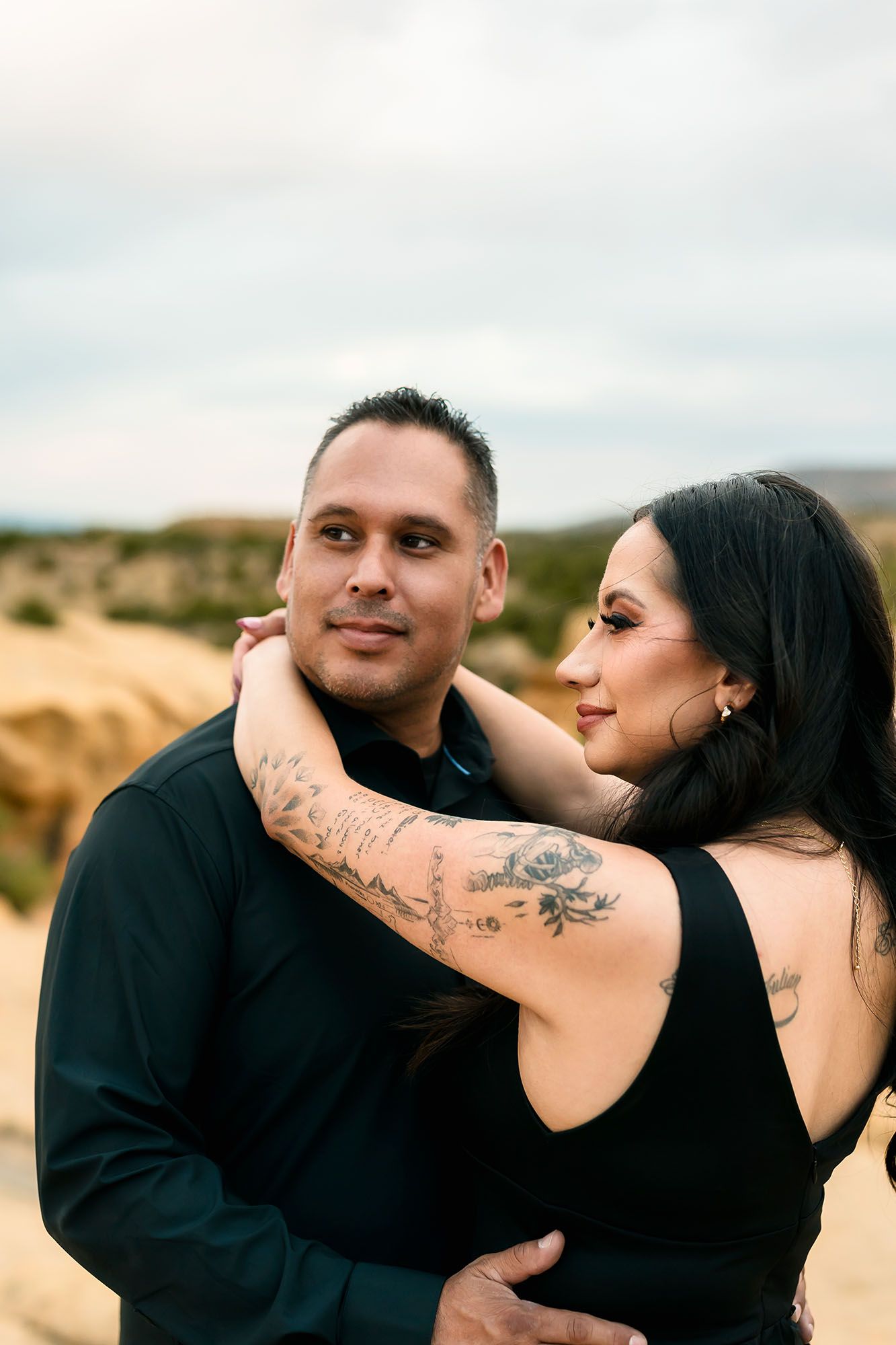 Tattooed woman in black dress embracing man from behind in romantic desert engagement portrait.