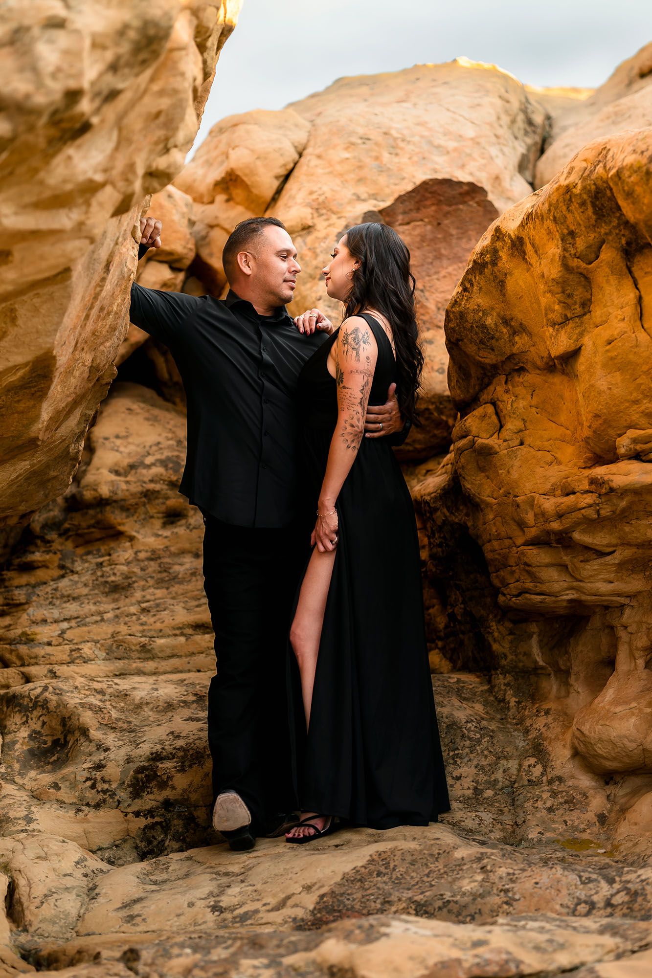 Smiling couple in black attire standing close together against warm golden rock formations outdoors.