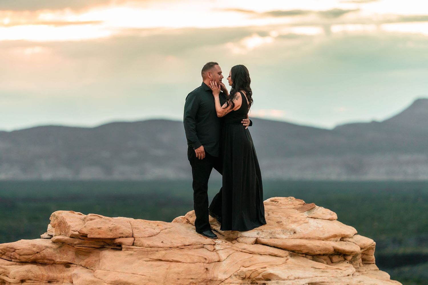 Couple in black attire sharing a romantic moment on a red rock cliff at sunset with mountain views.