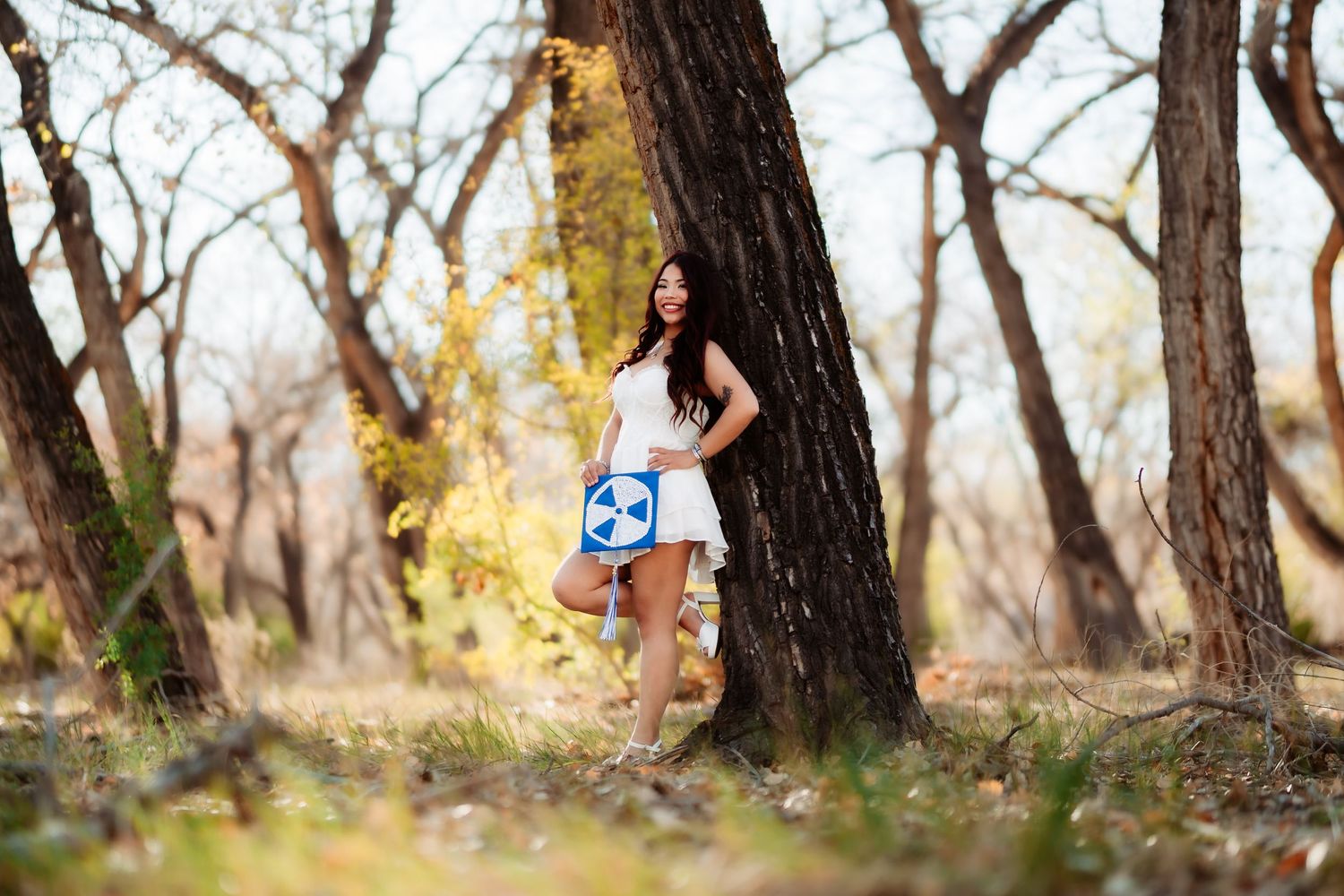 A woman poses gracefully in a natural outdoor setting with autumn trees and fallen leaves.