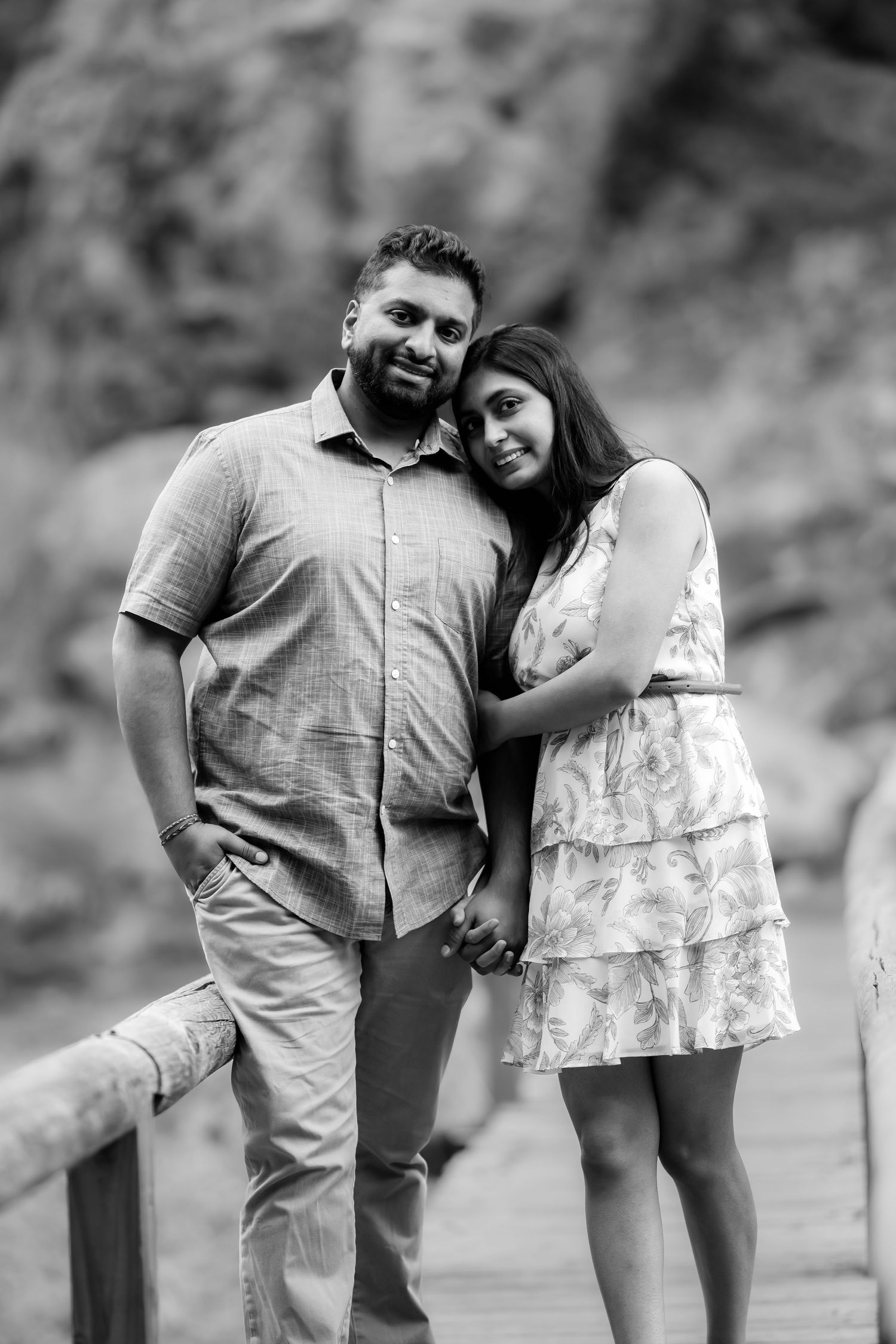 A couple in casual attire embraces while posing on a wooden walkway with rocky cliffs in the background.