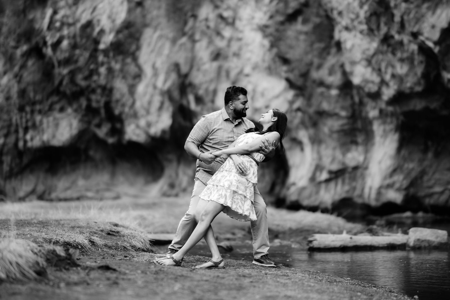 A black and white capture of a playful dance moment by the water with rocky cliffs in the background.