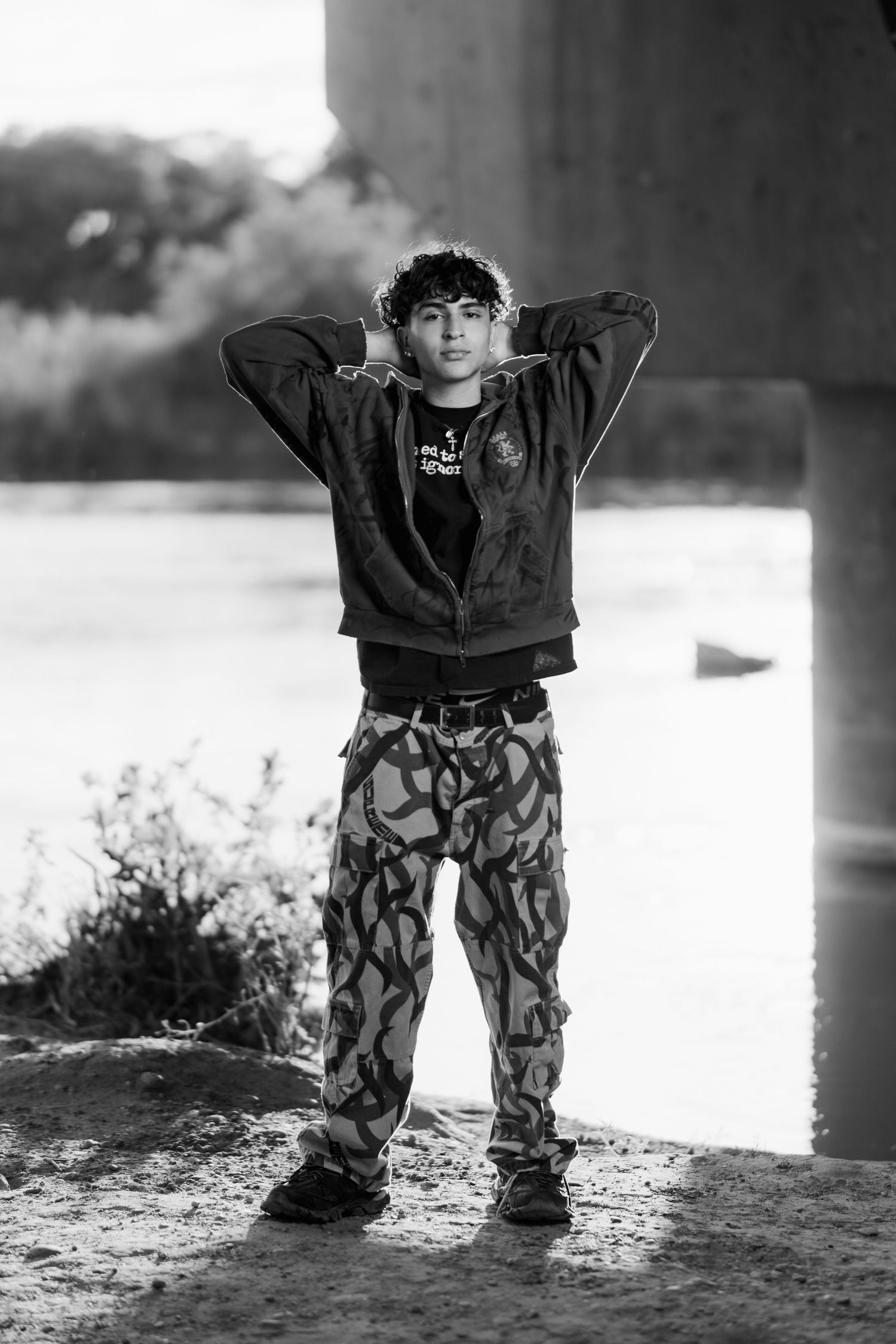 A person stands posing in patterned pants and a dark sweatshirt against a concrete wall in black and white.