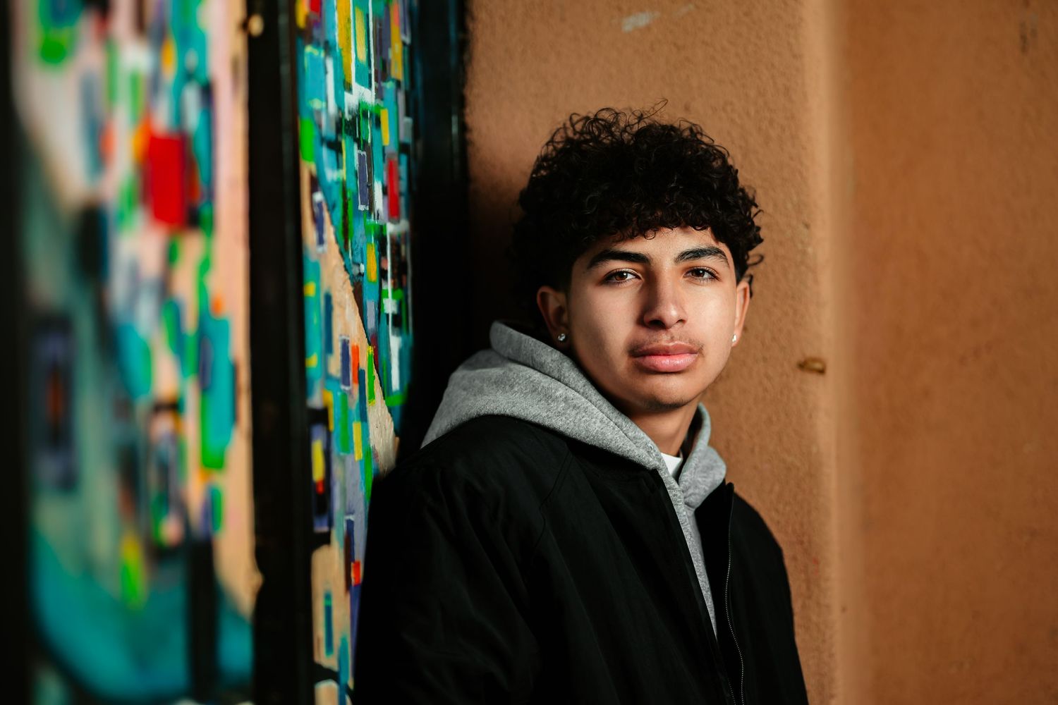 Young person in gray hoodie standing against textured wall with colorful graffiti art in background.