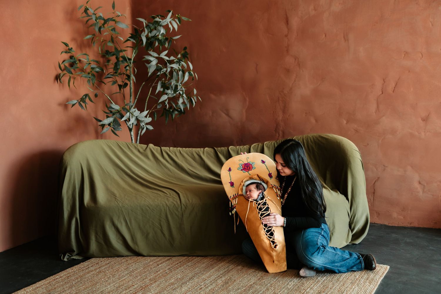 A greyhound dog rests on a green couch against a terracotta colored wall with potted plants in the background.