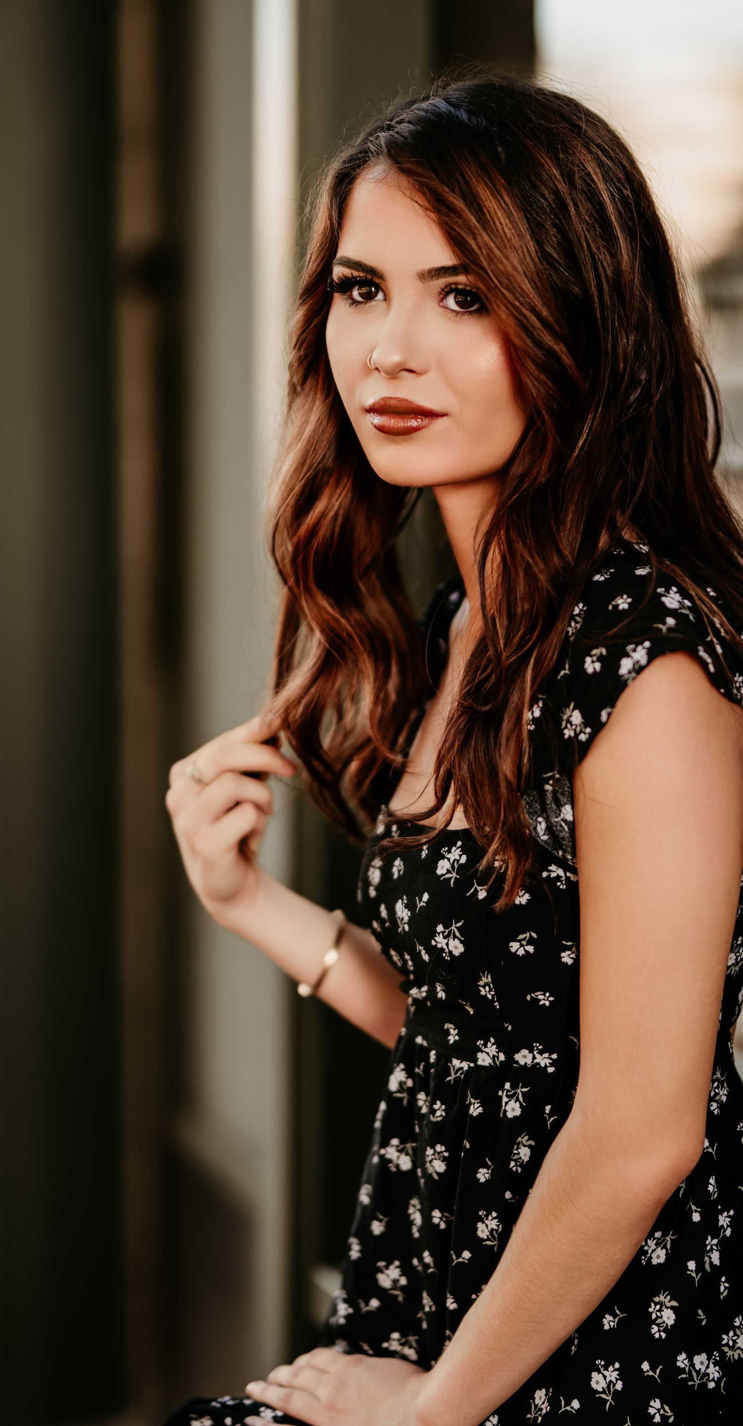 Woman in black floral dress poses against a blurred background with soft lighting and warm tones.