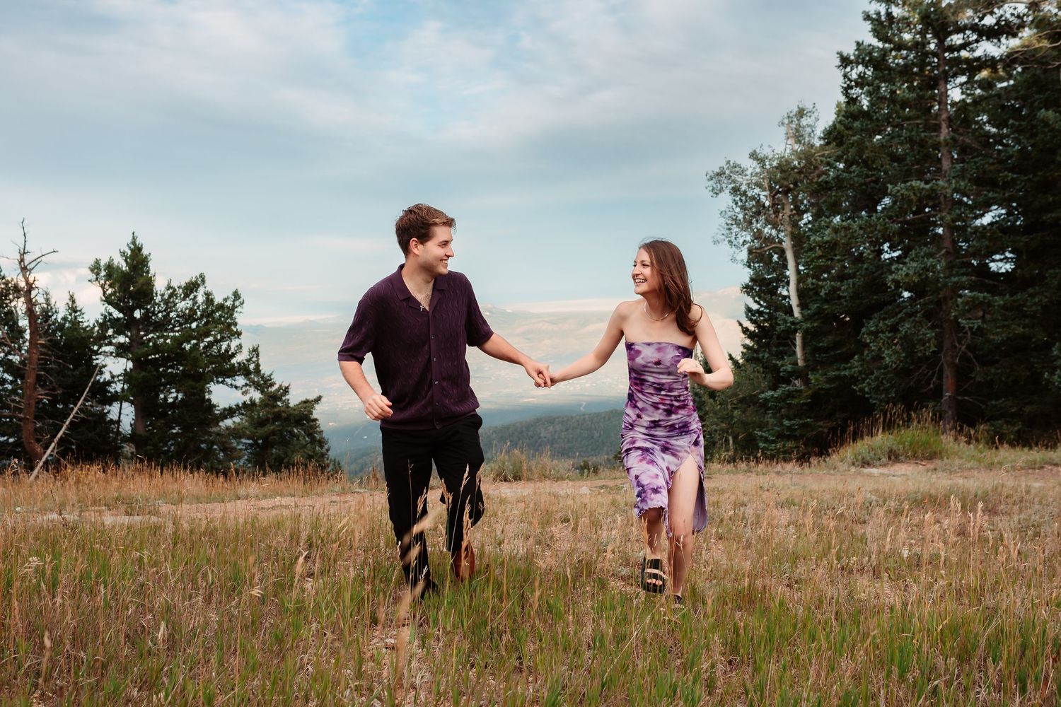 A joyful couple runs hand-in-hand through tall grass on a mountainside.