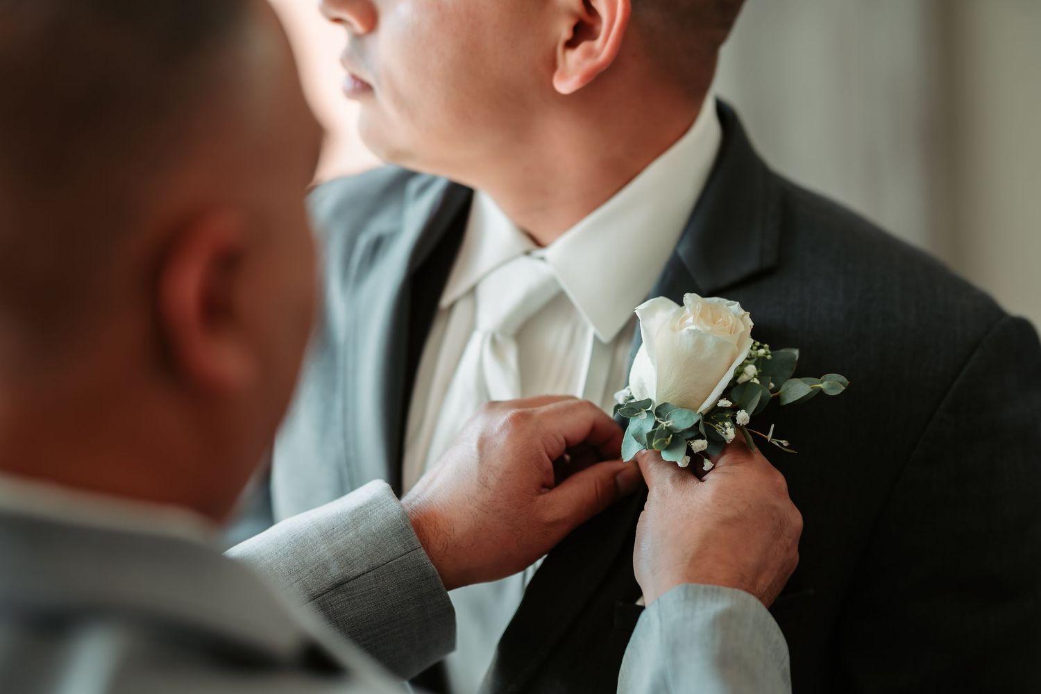 White floral boutonniere being pinned onto dark suit jacket during wedding preparations.