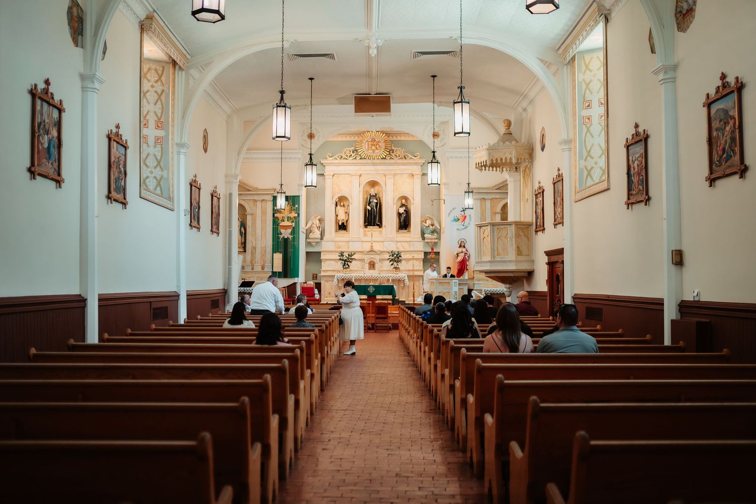 The interior of a traditional Catholic church with white walls, wooden pews, and arched ceilings illuminated by hanging lights.