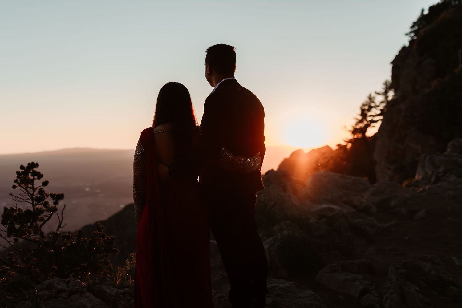 Silhouetted couple embracing against mountain landscape with dramatic sunset creating golden glow.