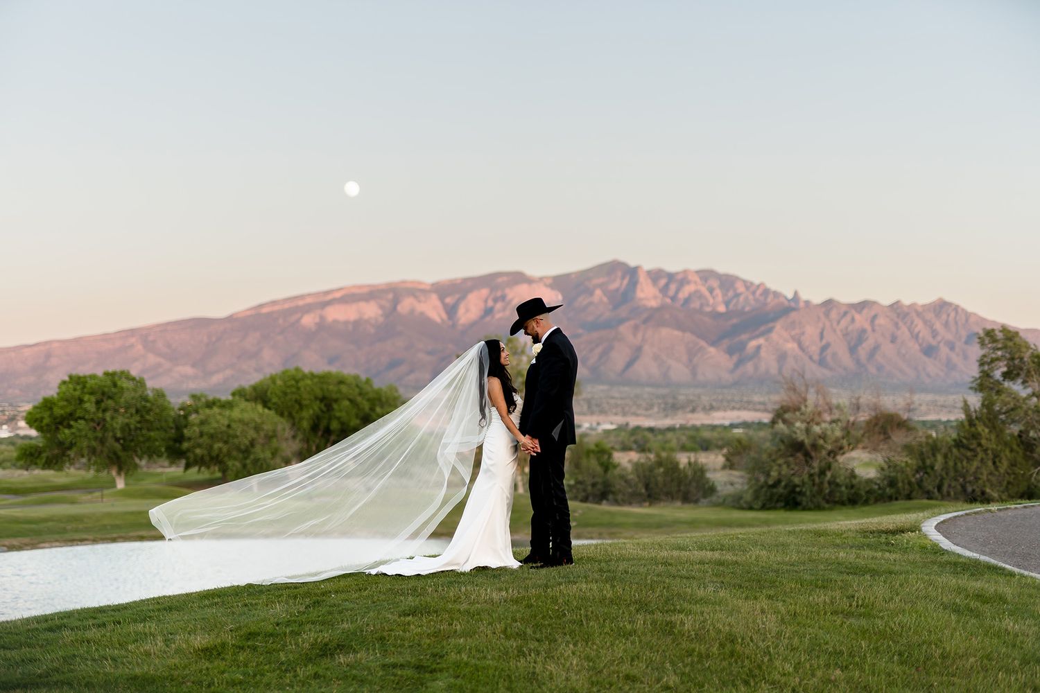 Bride and groom share romantic kiss at sunset with Sandia Mountains and full moon backdrop on lush green golf course.