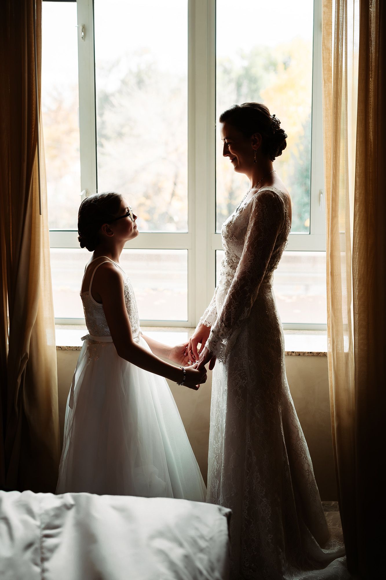 Bride and flower girl in white dresses holding hands by a sunlit window in a tender wedding moment.