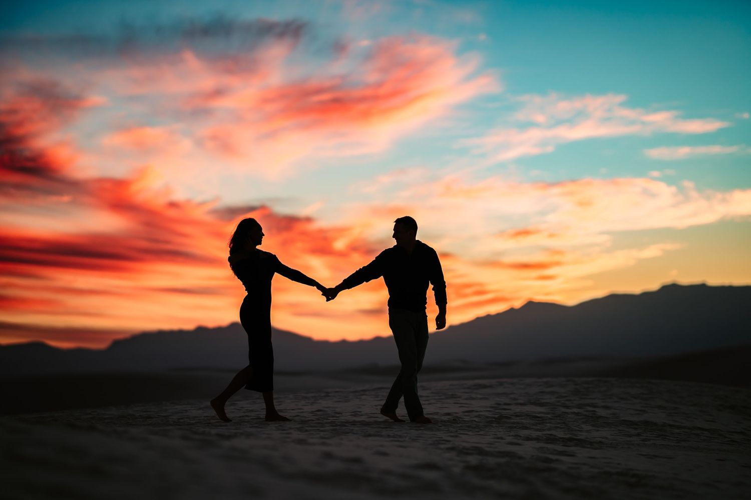 Two silhouettes hold hands against a dramatic orange and turquoise sunset sky over desert mountains.