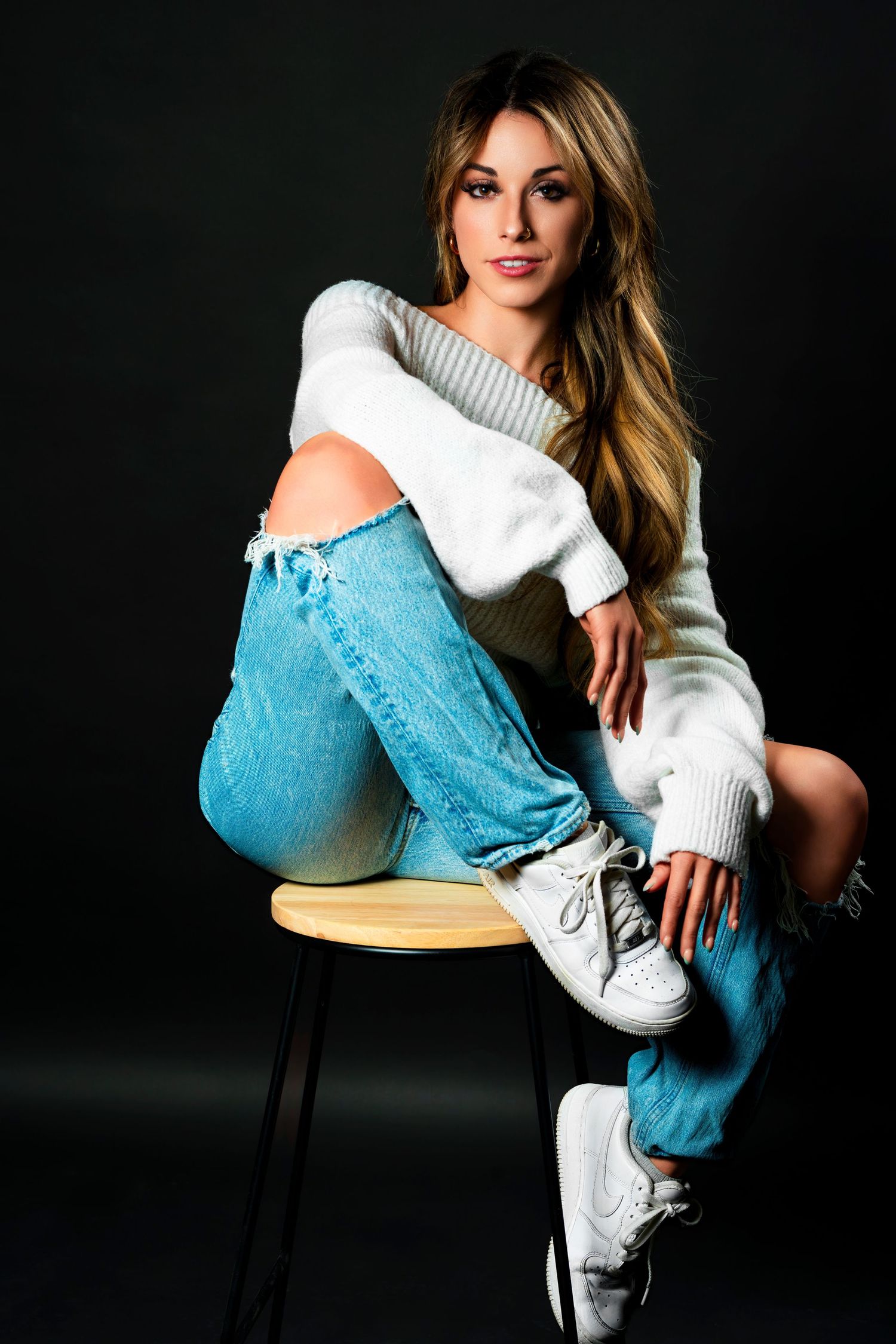 Fashion model in white sweater and light blue jeans poses on stool wearing white sneakers against dark background.