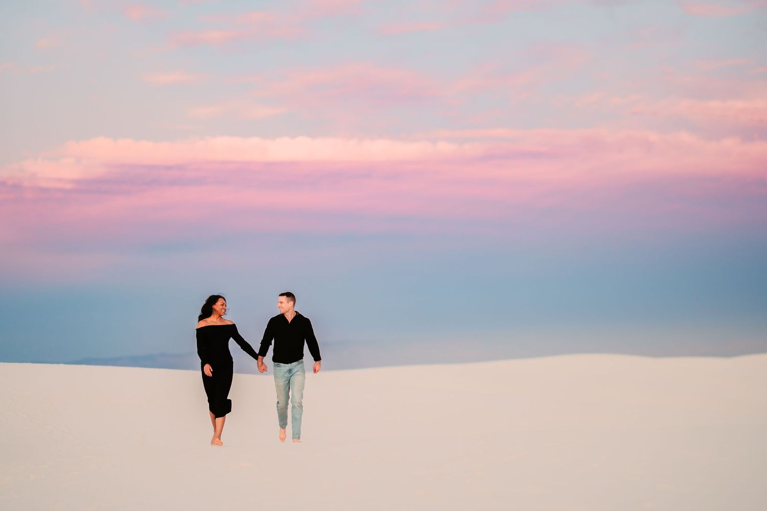 Silhouettes of people walking along a beach at sunset with pink and purple sky reflecting on the ocean.