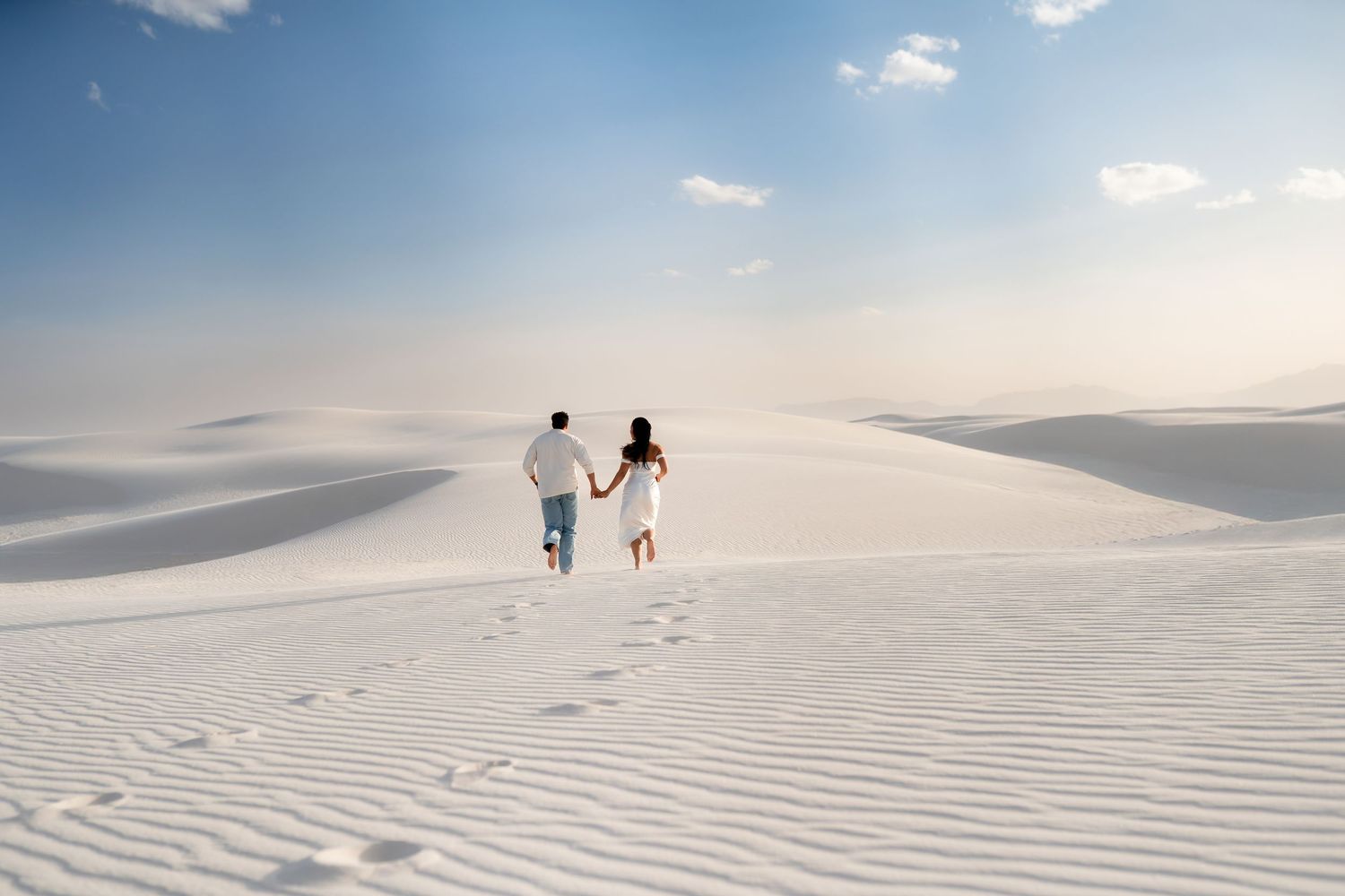 Two people walk across pristine white sand dunes under a soft cloudy sky, leaving footprints in their wake.