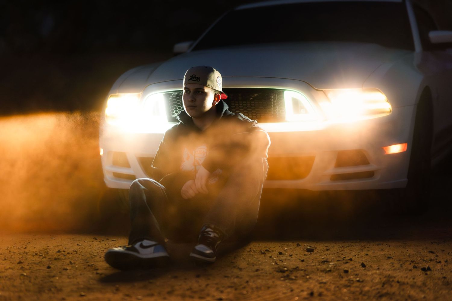 Young man sitting in front of glowing car headlights at night in a dramatic dusty outdoor scene.