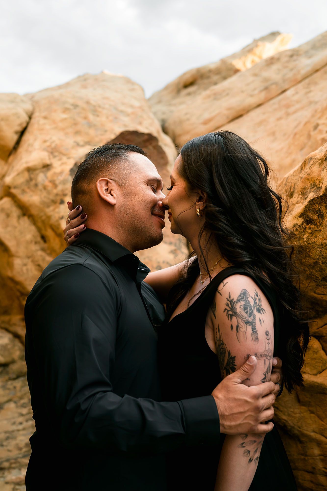 Romantic couple in black attire kissing against golden sandstone rock formation outdoors.