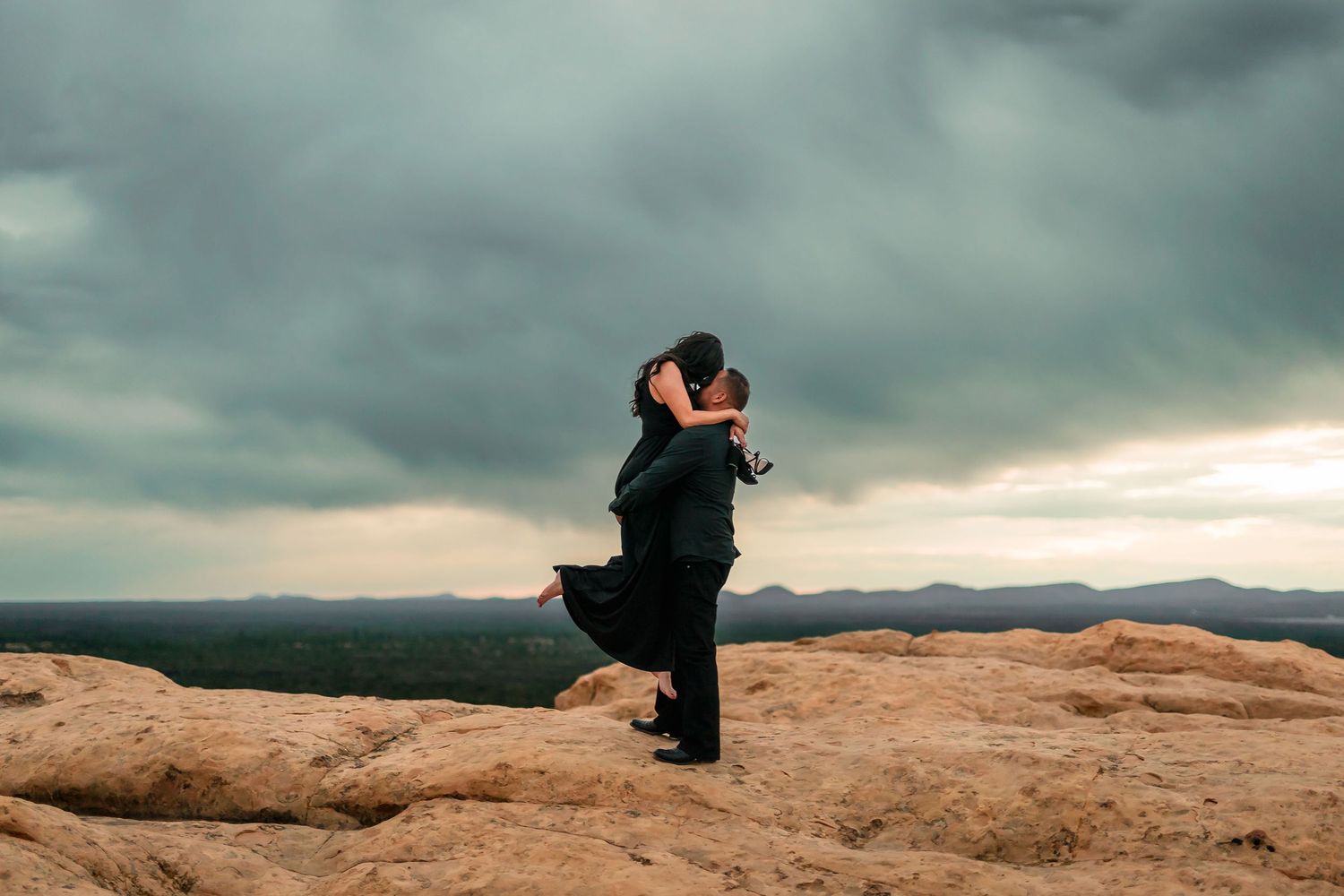 Man lifting woman in black dress during romantic engagement session on desert rock overlook.