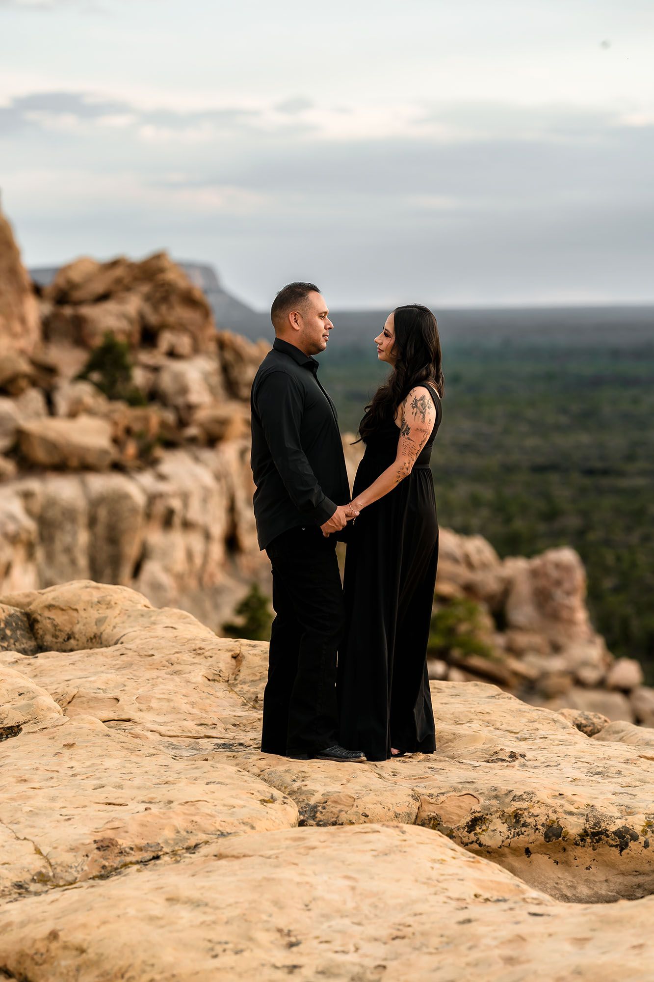 Couple holding hands in matching black outfits facing each other on rocky overlook with forest below.