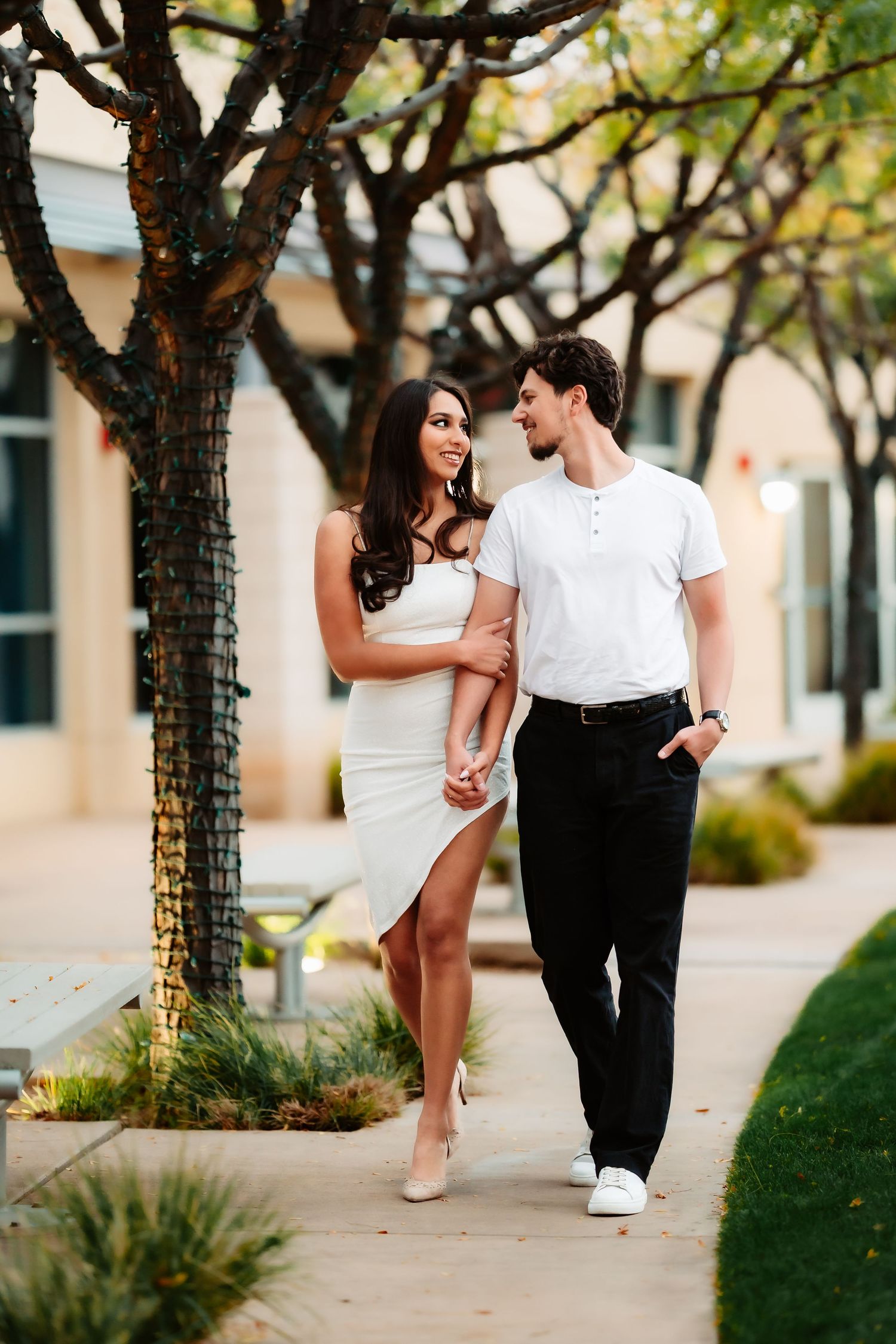 A couple in coordinated white and black outfits stroll together on a tree-lined pathway.