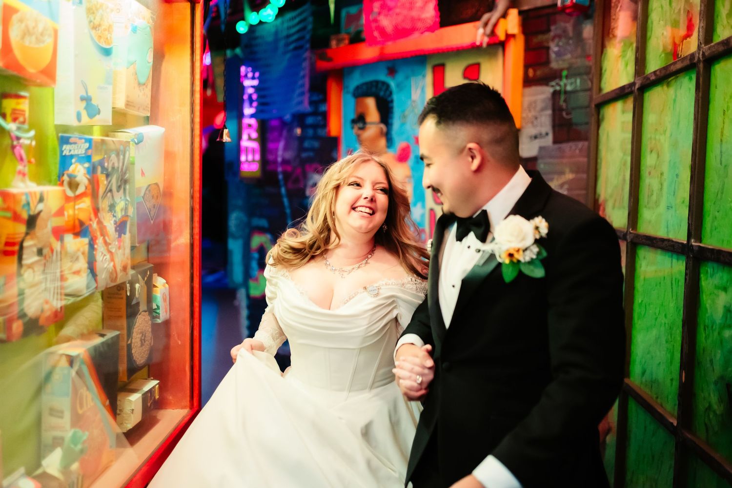 A couple in wedding attire shares an intimate moment in a colorfully lit urban alleyway.