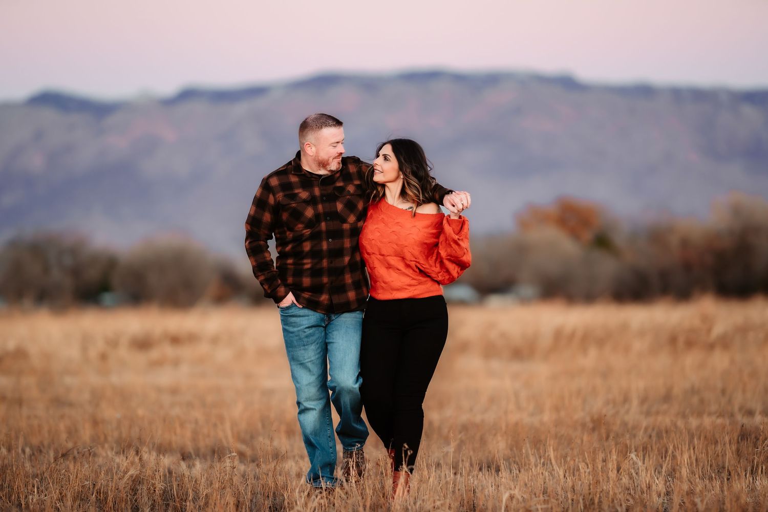 A couple walks together through a golden autumn field with mountains visible in the background at sunset.