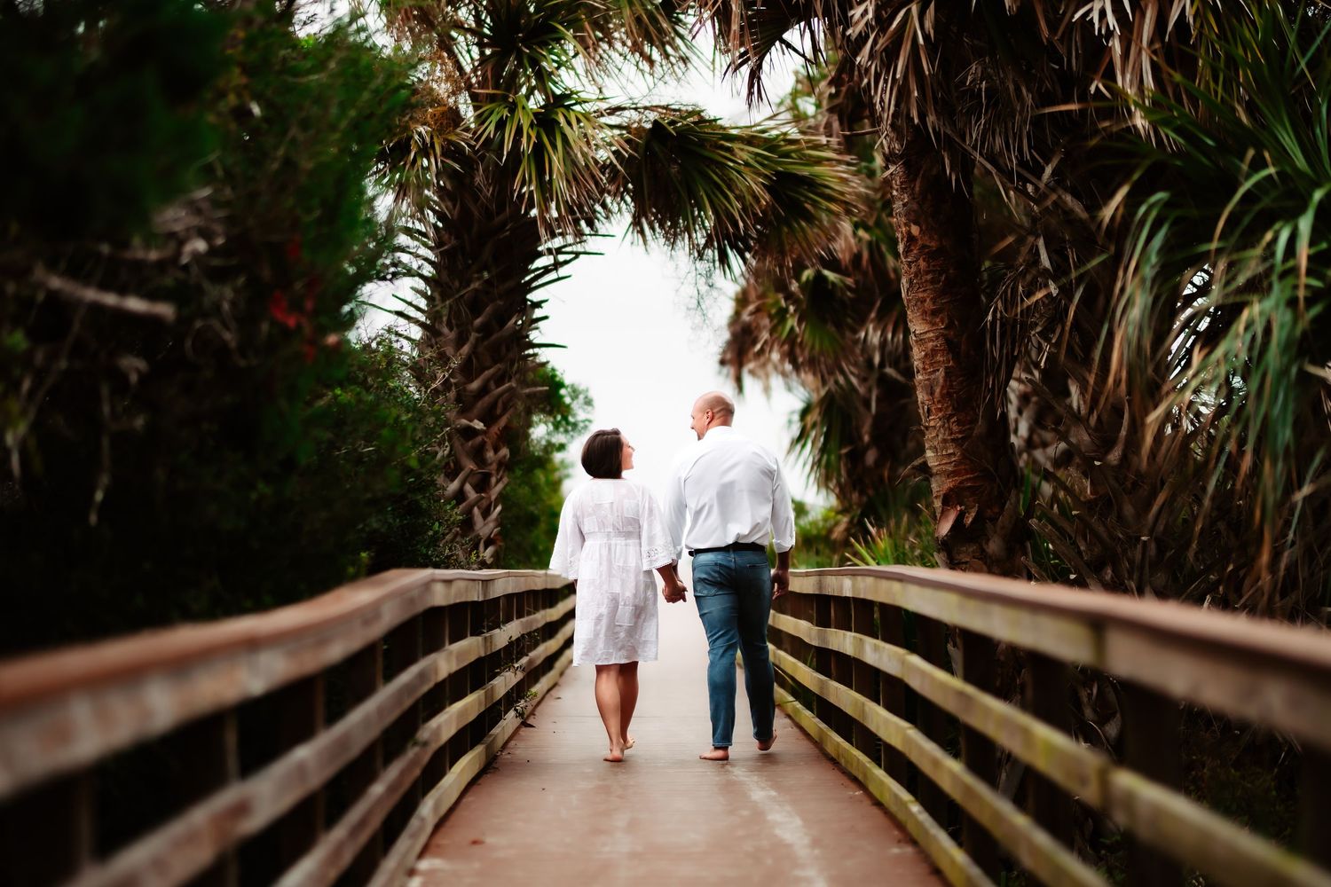 Two people walk hand in hand down a wooden boardwalk surrounded by lush tropical foliage.