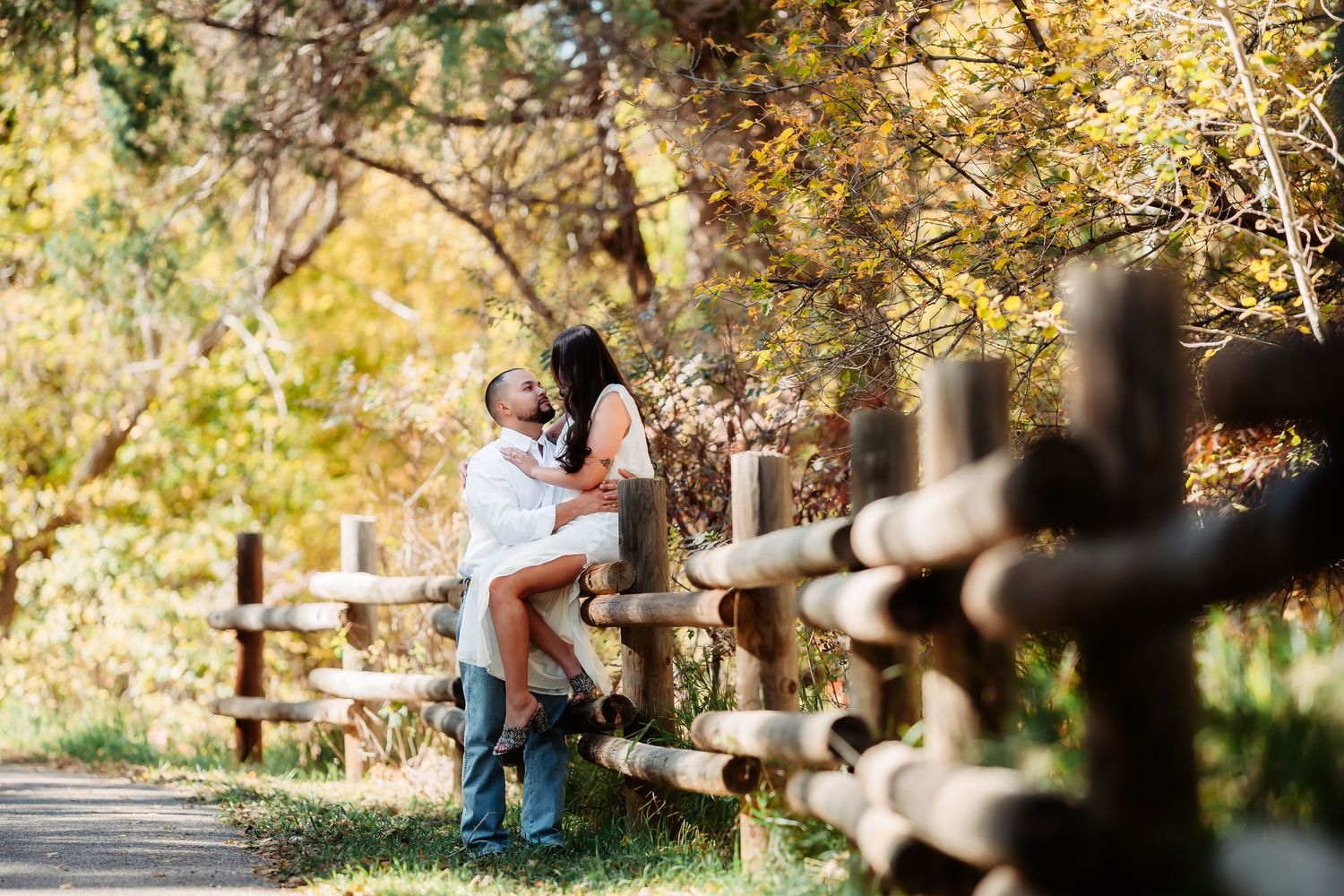 A couple sits together under autumn trees during a romantic outdoor photoshoot in a park setting.
