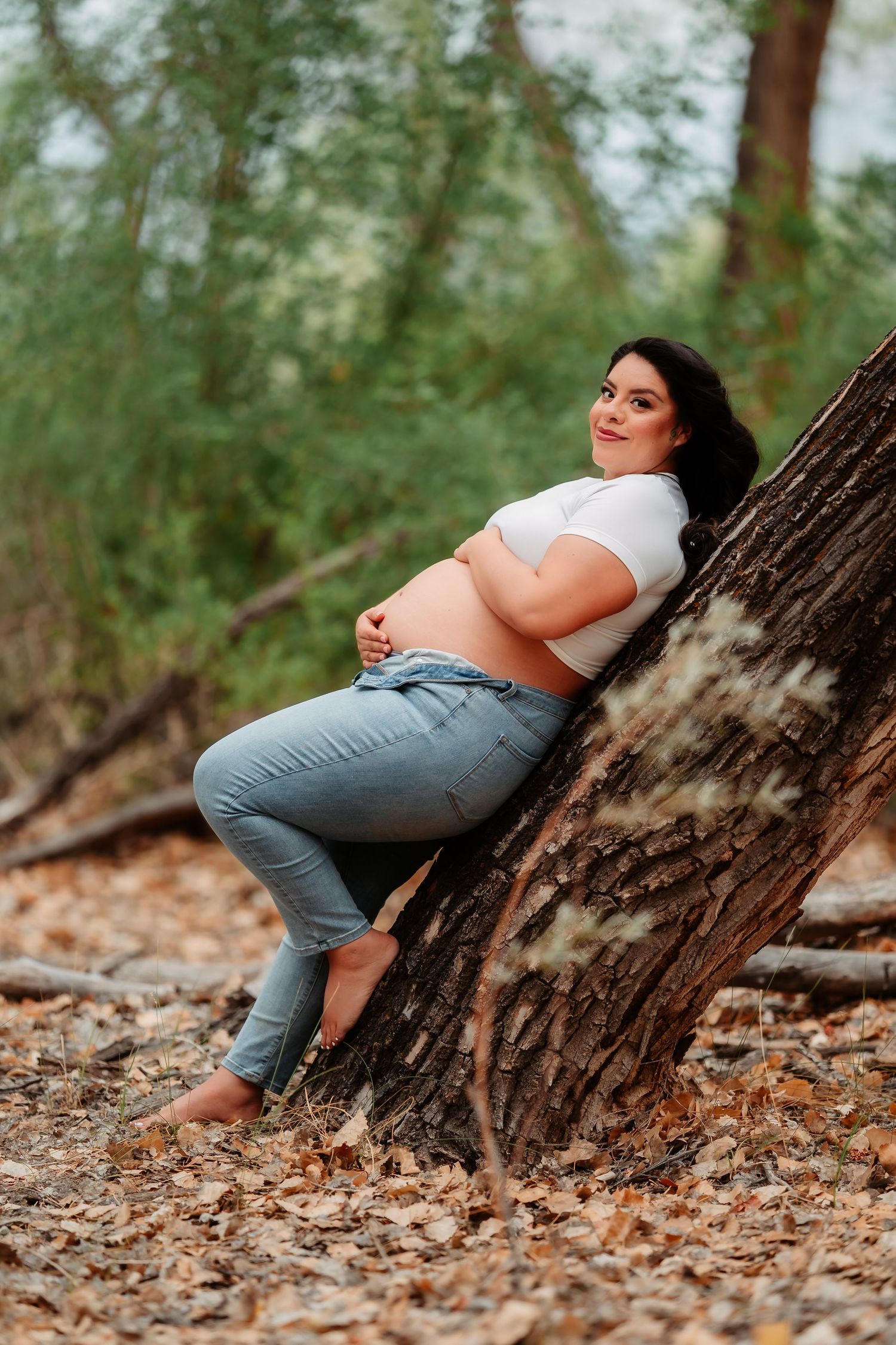 A person in casual attire lies comfortably against a tree trunk on a forest floor covered with autumn leaves.