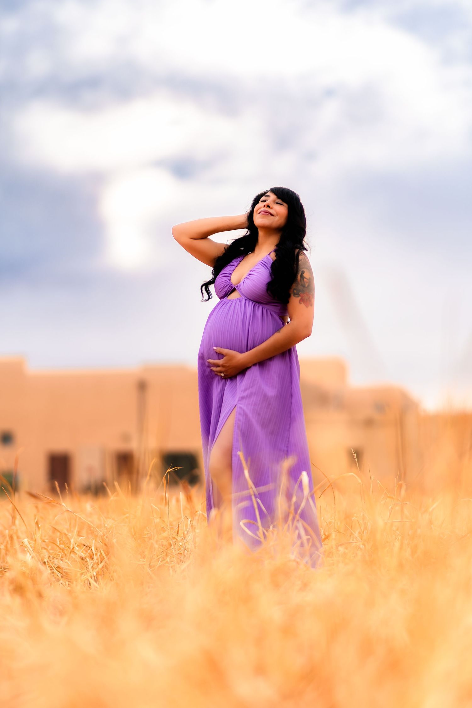 A pregnant woman in a flowing purple dress stands in a golden wheat field at sunset with a dreamy sky backdrop.