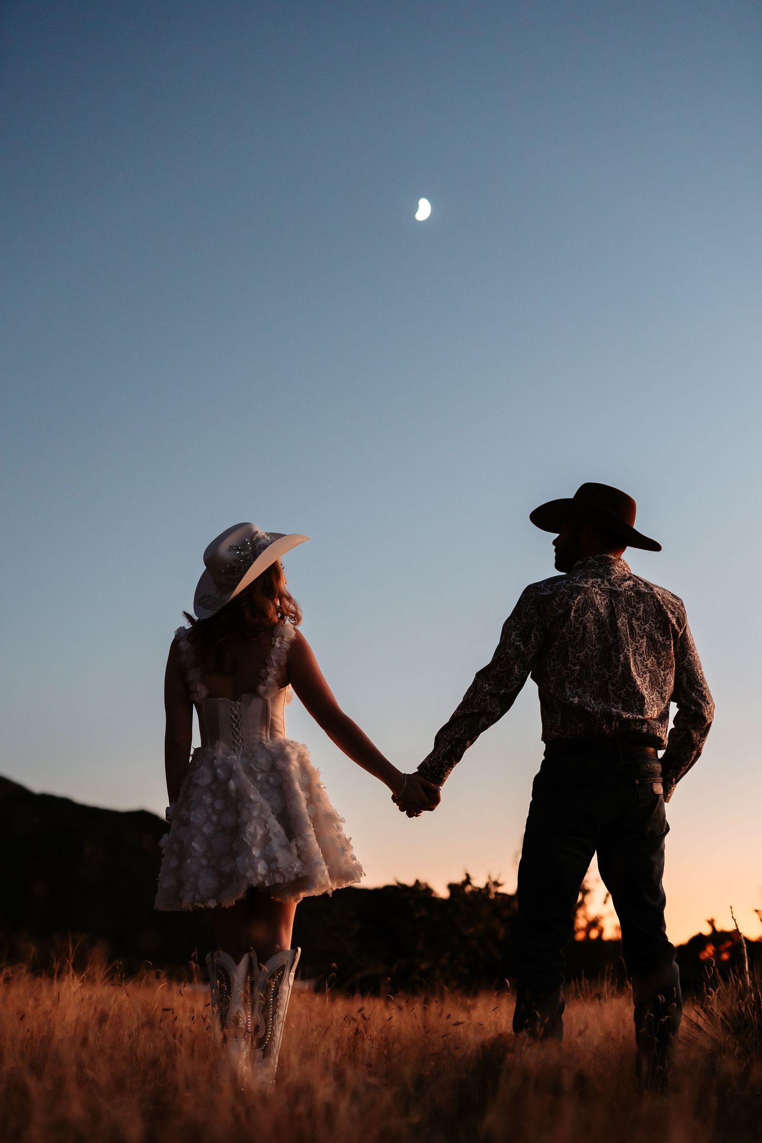 Silhouetted couple walking through a field at sunset, holding hands and leaning towards each other in a romantic pose.