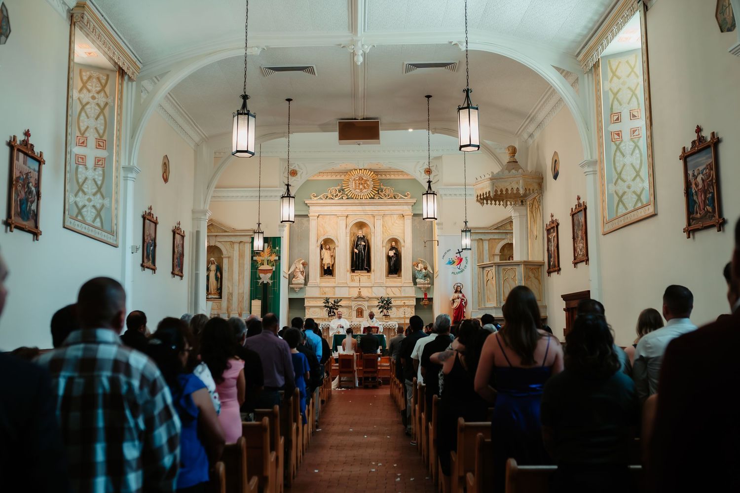 People gather inside a traditional Catholic church with white arched ceilings and pendant lights during a service.