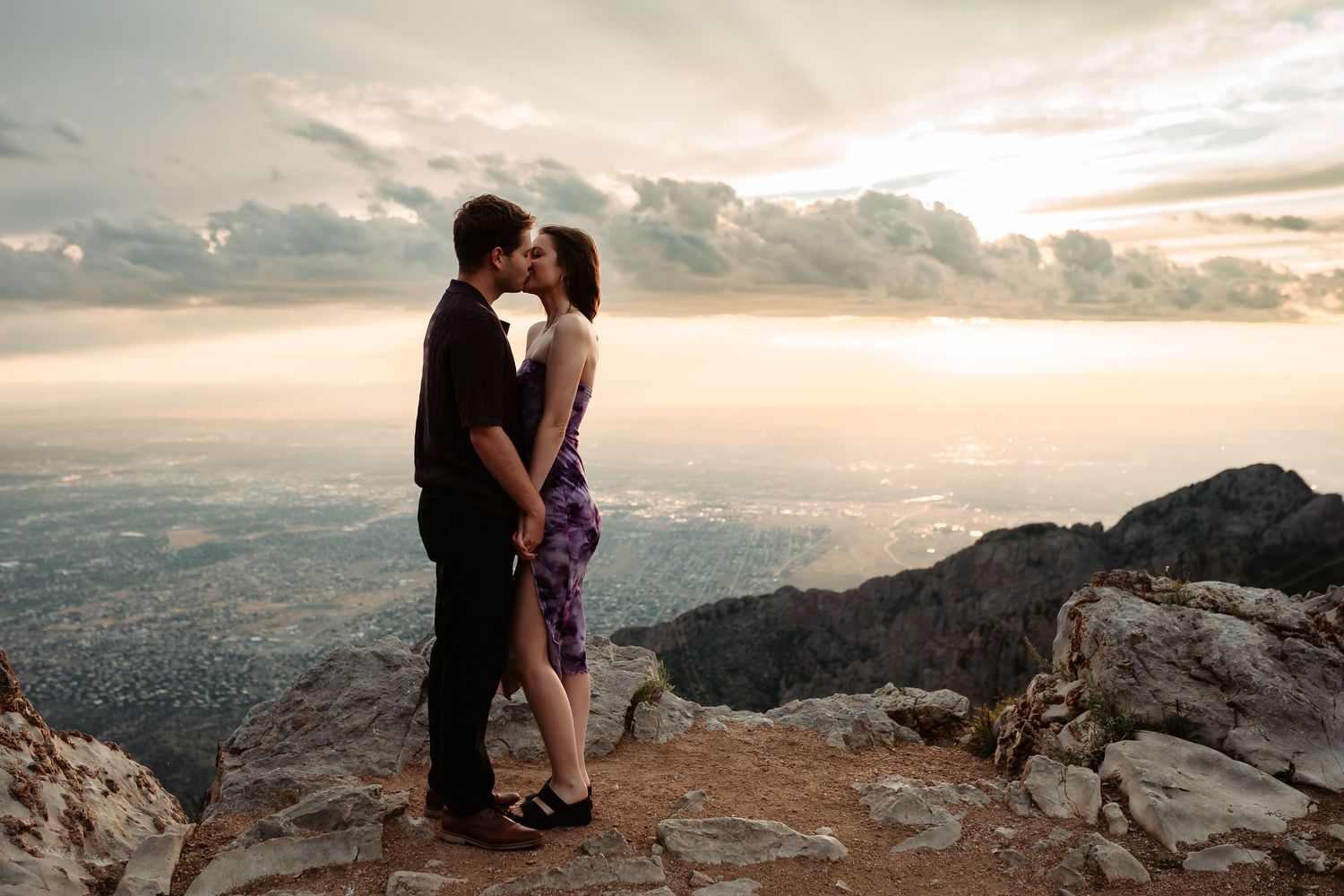 Couple shares romantic moment on mountain cliff overlook at sunset with dramatic cloudy sky in background.