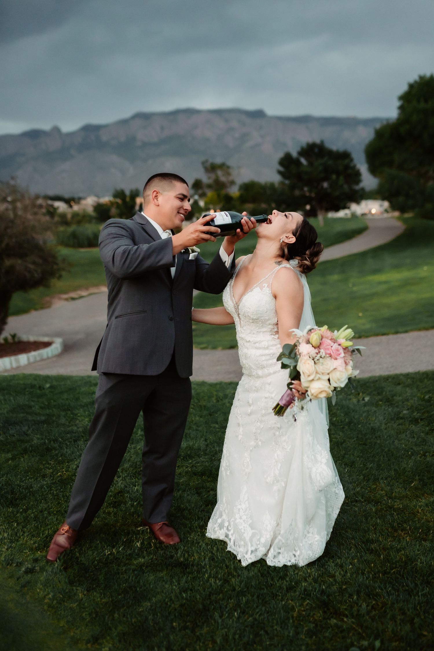 Playful wedding couple taking celebratory shots on grassy hillside.