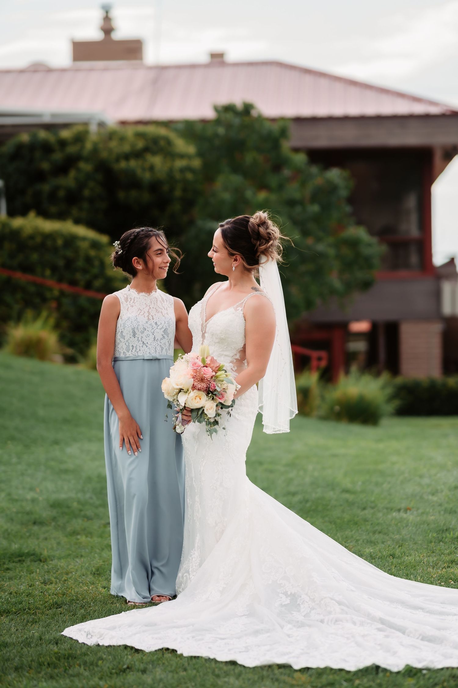 Two figures in light blue bridesmaid dresses and white wedding gown pose together on a grassy lawn near a building.