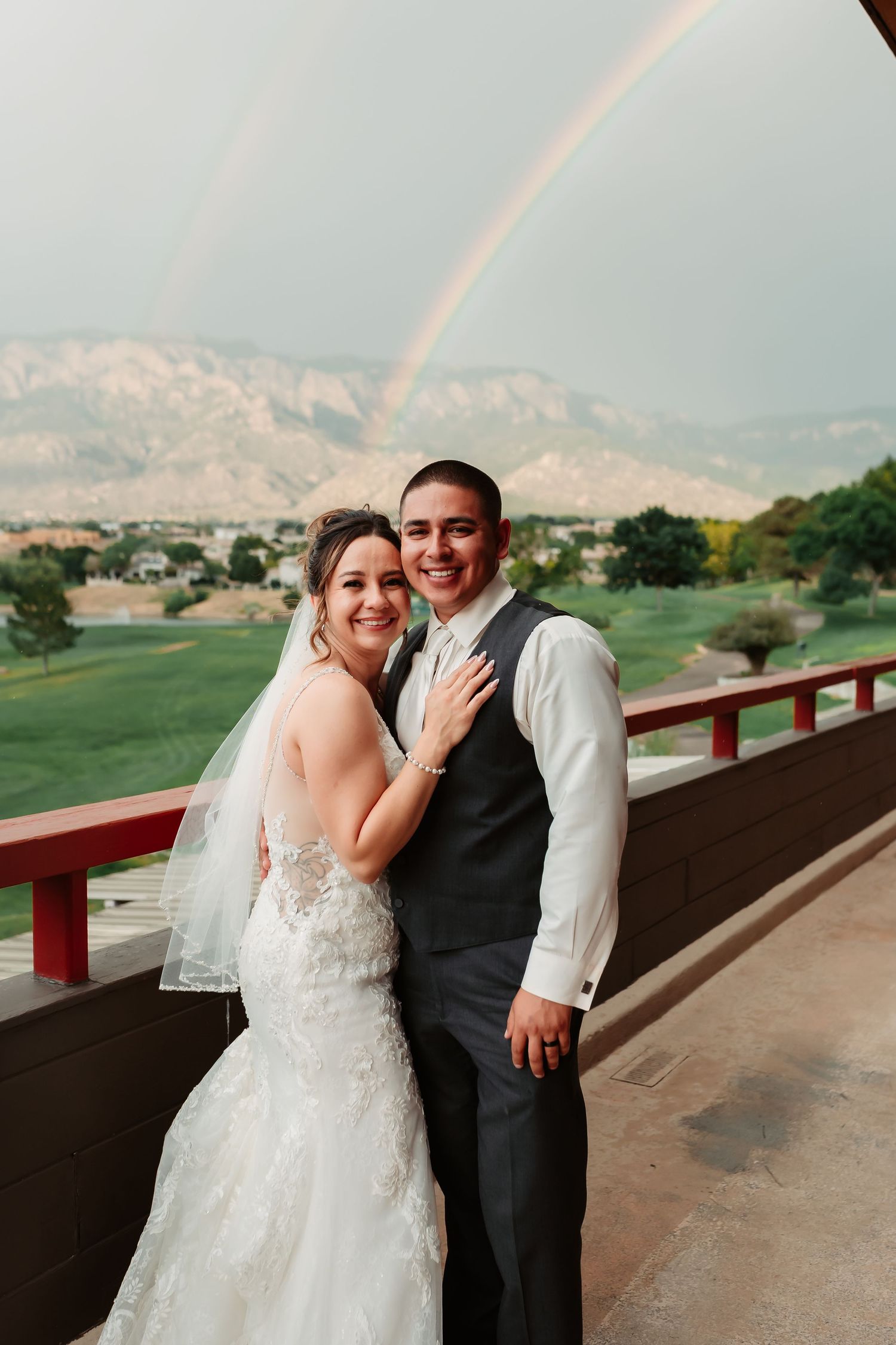 A joyful wedding couple shares a romantic moment on a balcony overlooking mountains and golf course at sunset.